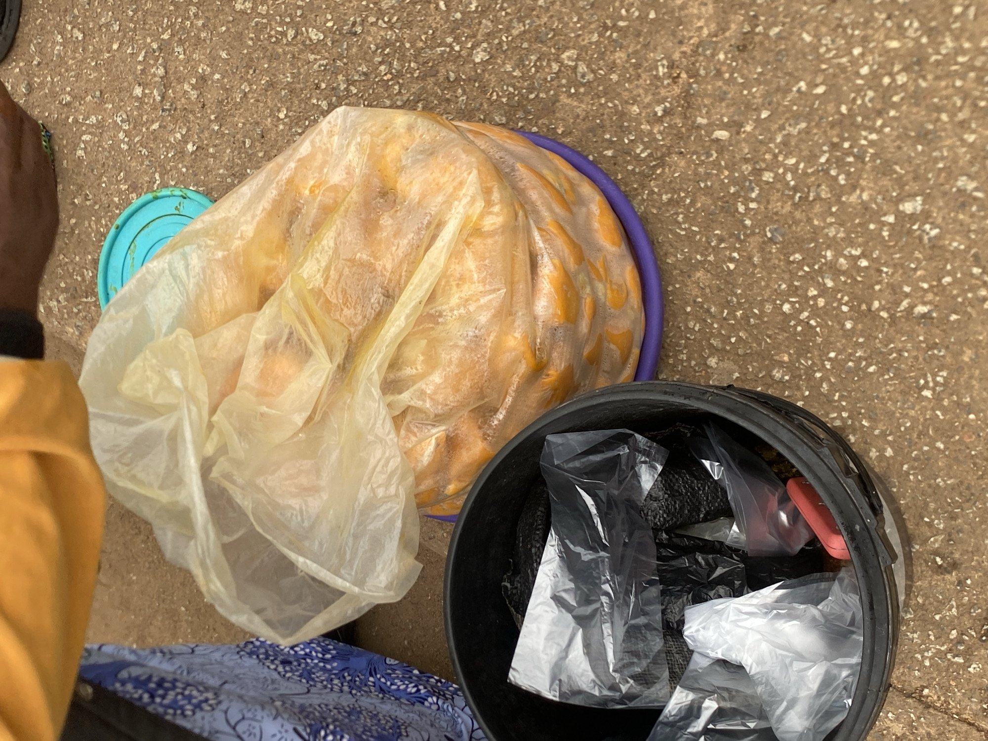 In Bukuru, Nigeria, on a rough, granular ground surface, a black plastic bucket is positioned to the left, containing several plastic bags, some clear and others black. Inside the bucket, a small red object is visible among the plastic bags. To the right of the bucket, a large, translucent yellow plastic bag is filled with a bulk of what appear to be yellow fruits, stacked on top of a purple plastic lid. A portion of a person's hand, wearing a black glove, is partially visible reaching towards the yellow bag, and the person is dressed in an orange garment. A blue plastic lid is also partially visible near the yellow bag.