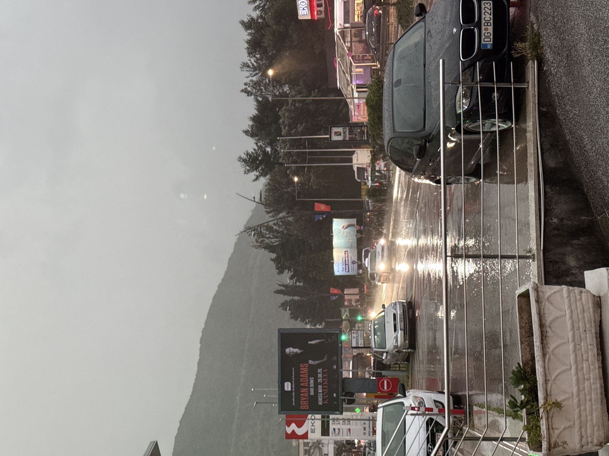 This media depicts a street scene in Budva, Montenegro, under overcast and rainy conditions. The wet asphalt reflects the bright headlights of passing vehicles and illuminated streetlights, suggesting it might be evening or a heavily clouded day. Mountains shrouded in mist or low clouds are visible in the background.
The foreground features a black BMW sedan parked on the side of the road, partially obscured by a metal railing. Its license plate, "DG BC223," is clearly visible. Raindrops are evident on the car's surface. To the right, a concrete planter with sparse greenery sits beside the railing.
Along the road, several cars are visible, some with their headlights on, moving through the wet conditions. A white van or car is prominent behind the railing further down the street. Buildings, likely commercial, line the road with glowing signs.
Notable signs include an "EKO" logo, possibly for a gas station or shop, and a large billboard featuring "BRYAN ADAMS" for a "BARE BONES" concert scheduled for "26.06.26." in "HERCEG NOVI KANLIKULA," identifying a specific Montenegrin location. Above this, another billboard contains text including "GINEKOLOG" and "Val biznis. Naša podrška." A "NAMOS" sign is also visible further down the street. The overall atmosphere is one of an urban area during inclement weather.