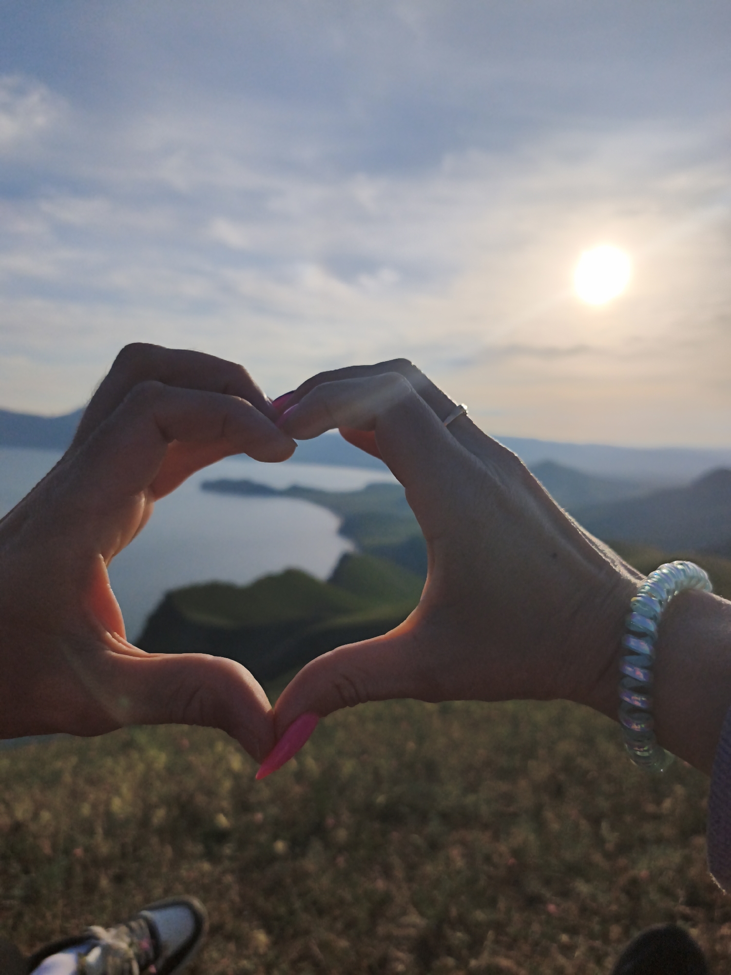 A person stands on a grassy hill during a sunny day, forming a heart shape with their hands. Through the heart shape, a lake, distant hills, and a bright sun are visible. The person wears a ring on their right hand and a spiral-shaped bracelet on their right wrist. A shoe is visible in the lower left corner. The location is not identifiable as Ordzhonikidze, Ukraine from the given information.