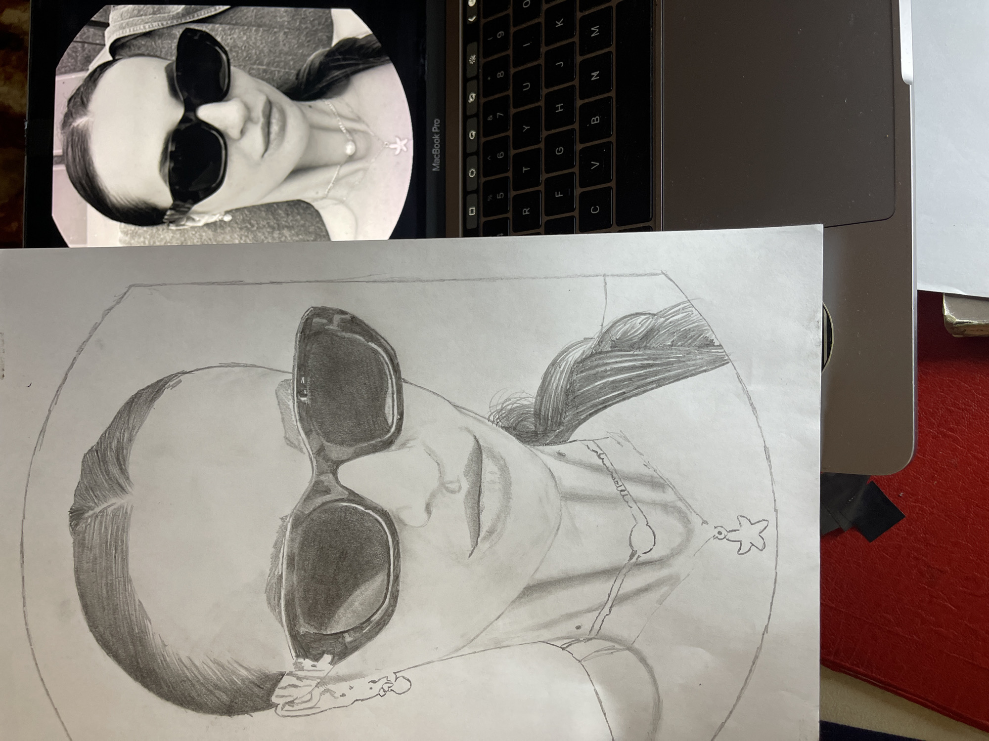 The image displays a pencil drawing of a woman wearing sunglasses, alongside the photograph it is based on, both placed on top of a partially visible laptop. The drawing is a sketch, with shading and outlines visible, and includes details like her hair, sunglasses, and necklace. The photograph on the laptop screen shows the same subject in black and white, providing a reference for the drawing. The laptop's keyboard is partially visible in the top right corner of the frame. The scene appears to be indoors, with other items like papers and a red-covered object visible in the background. The location of the city of Jos, Nigeria is not present or discernible in the image.