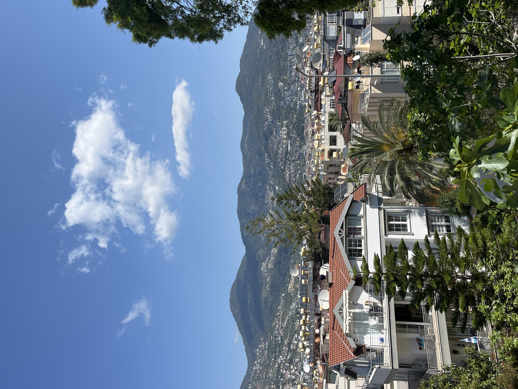 The image shows a view in Alanya, Turkey. A mountain rises in the background, densely covered with vegetation and dotted with buildings. In the foreground are white buildings with red-tiled roofs, trees, and satellite dishes. The sky is blue with scattered white clouds. There is a palm tree among other leafy trees. The buildings are densely packed together.