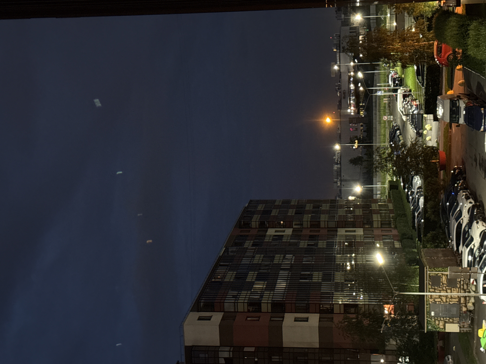 The image shows a view from a window at dusk or night in an urban environment. A tall, multi-story apartment building with a facade featuring colorful vertical elements and numerous windows dominates the left side of the frame. In front of the building, a parking lot is visible, filled with numerous cars, mostly sedans and SUVs, parked in rows. Streetlights illuminate the scene, casting a warm glow. Several trees with dark foliage are scattered throughout the parking area and along the side of the building. In the distance, other buildings and what appears to be a commercial establishment with illuminated signs are visible. The sky is a deep blue, with some faint, blurry light phenomena that could be reflections or atmospheric effects. The overall impression is one of a residential area at the end of the day, with artificial lights starting to take over from the fading natural light.