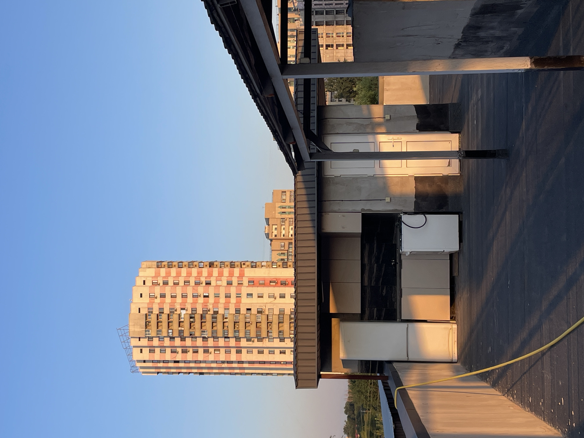 The image captures an outdoor scene on a rooftop with a clear blue sky overhead. In the background, a tall, pink and white residential building stands prominently, its facade punctuated by numerous windows and balconies. To the left, the edge of the rooftop features a low concrete wall with a yellow hose draped over it. In the mid-ground, a series of outdoor kitchen structures are visible, including white appliances, cabinets with dark countertops, and white doors. The area is illuminated by the warm glow of sunset, casting long shadows across the dark grey rooftop surface. The architecture suggests an urban setting, likely in Almaty, Kazakhstan, characterized by high-rise residential buildings and the functional design of rooftop amenities. No people are present in the image.