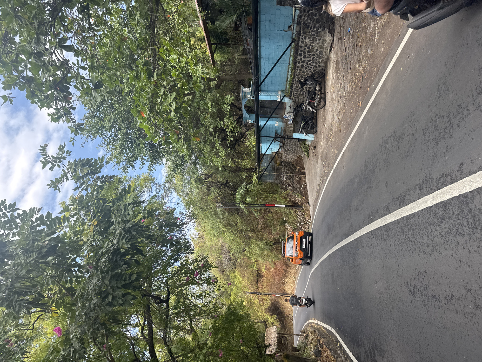 The image shows a road winding through a lush, tropical environment, likely in Abang, Indonesia. In the foreground, the asphalt road is bordered by white lines and trees with dense green foliage overhead. A person on a scooter rides ahead on the left side of the road, followed by an orange car. To the right of the road, a bright blue wall with stone foundations encloses a property with several parked motorcycles. Further back, more trees and vegetation cover the hilly terrain under a partly cloudy sky. The overall scene suggests a scenic route in a rural or semi-rural area.