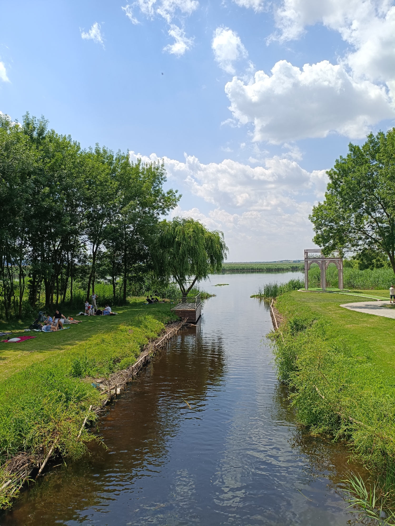 A scenic view of a canal leading to a lake in Comana, Romania, features a lush green park on the left bank where several people are picnicking and relaxing on the grass under the shade of tall trees. A weeping willow tree droops over the water, near a wooden pier. On the right bank, a paved path winds through the grass towards an ornamental archway. Further in the distance, reeds line the lake's edge, and the sky is adorned with fluffy white clouds against a bright blue backdrop.
