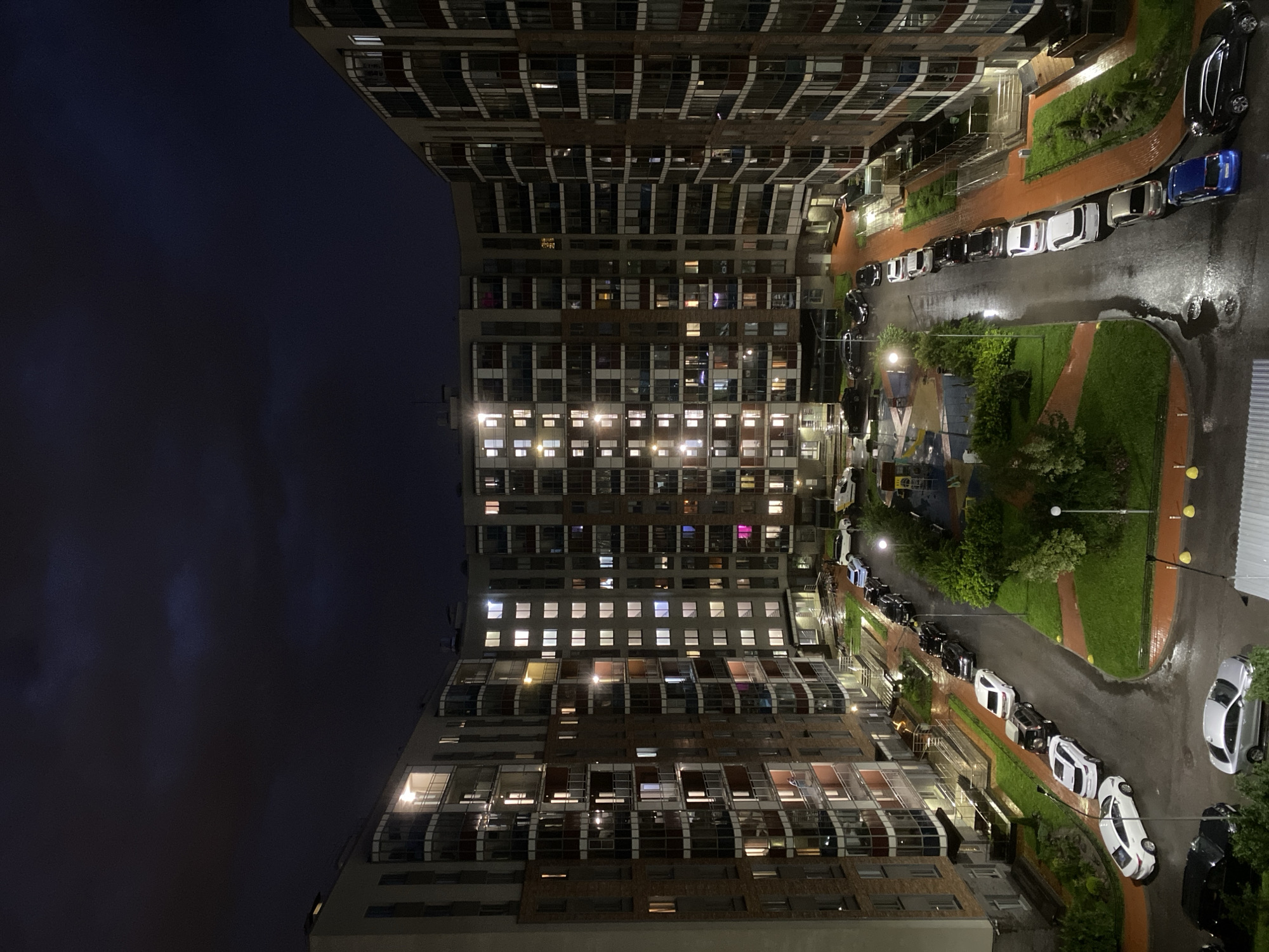 An aerial, night-time photograph depicts a modern, U-shaped residential apartment complex with multiple stories and numerous lit windows. The courtyard area features a playground with slides and swings, surrounded by green grass, paved walkways, and a wet asphalt road. Several cars are parked along the road and in designated spots. The sky above is dark with visible clouds, suggesting a possibly overcast or stormy night. The buildings are characterized by brick and glass facades, with balconies visible on several floors. The scene is set in Ladozhskoe Ozero, Russia.