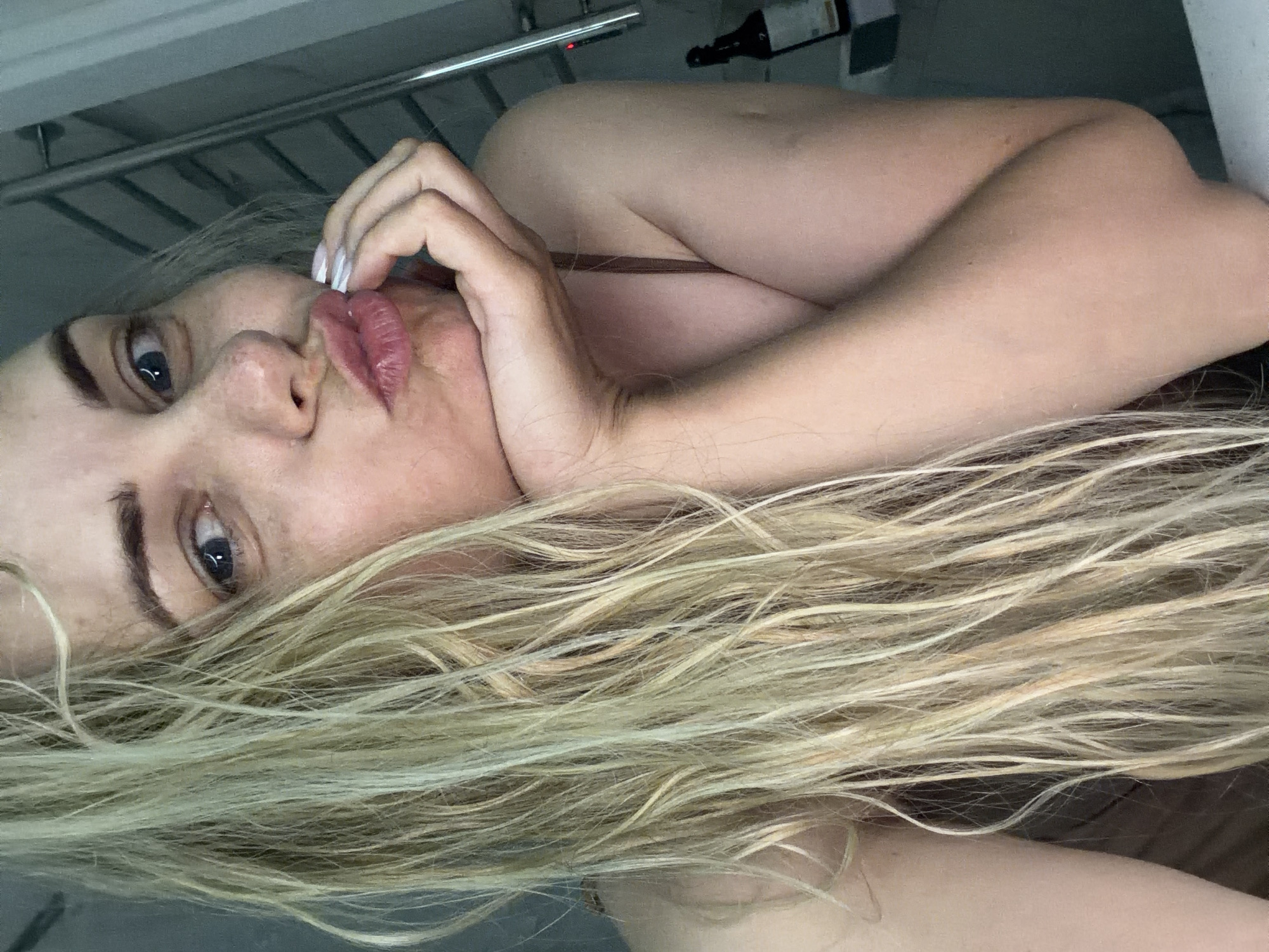 A light-skinned woman with blonde hair lies on her side, looking toward the camera. She has blue eyes and is pouting her lips while holding her hand to her face. Part of a silver metal shelf structure is visible in the upper left corner. A dark bottle sits on a shelf in the upper center of the image. The location is not evident in the image.