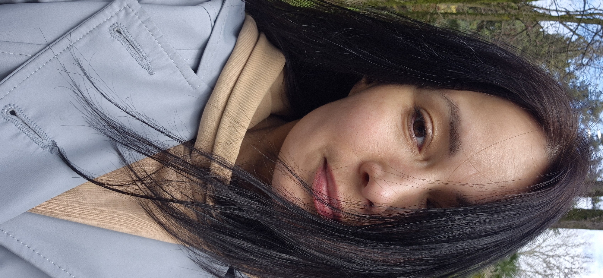 A close-up, slightly rotated image shows a woman with dark hair, fair skin, and brown eyes. She is wearing a light blue coat and a beige hoodie. Wisps of her hair are across her face. The background shows trees and a blue sky. The location is unknown and there is no basis for presuming it is Sochi, Russia.