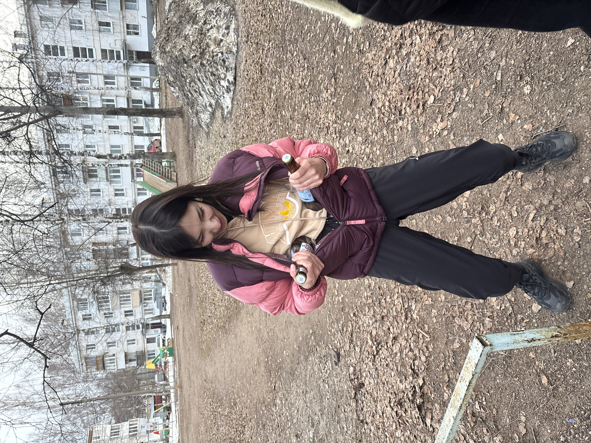 The image shows a young woman outdoors, holding two bottles of what appears to be beer. She is standing on a ground covered with dry leaves and small twigs. She is wearing a pink and maroon jacket, a beige shirt, and dark-colored sweatpants. She is also wearing dark sneakers. In the background, there is a multi-story apartment building and a playground. The location is likely in a residential area of the city of Chaikovsky, Russia, though this cannot be definitively determined from the image alone.