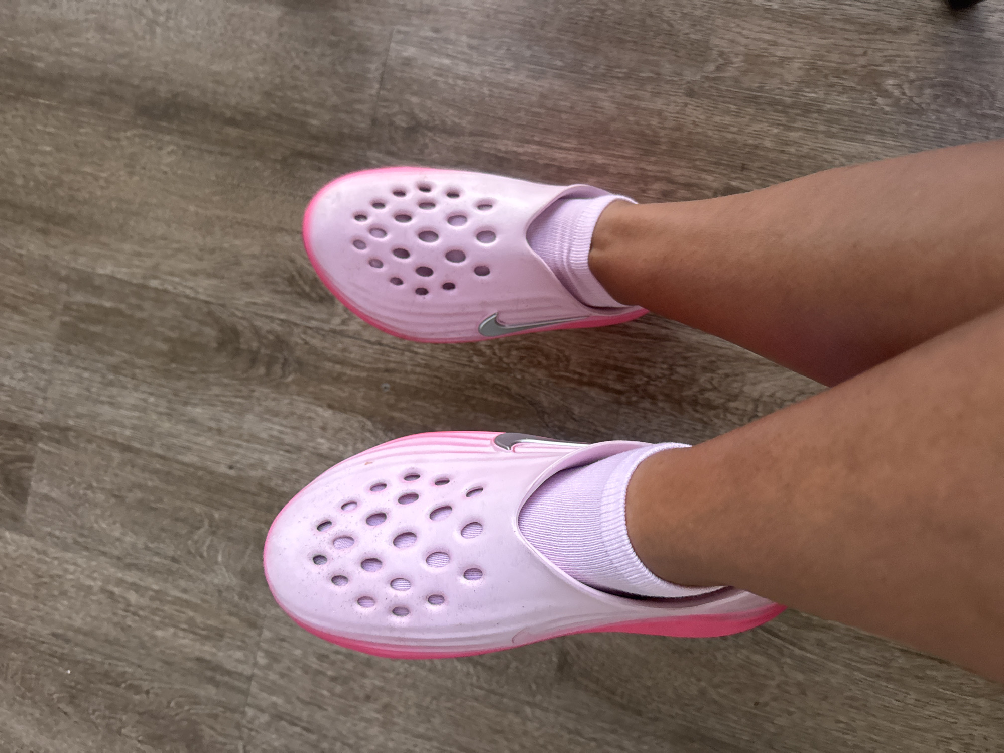 A pair of light purple clogs with pink soles and a silver Nike logo are worn by a person standing on a wooden floor. The clogs have multiple circular perforations on the top. The person is also wearing light purple ankle socks. The image is a close-up shot of the person's feet and lower legs. The background is a wooden floor with visible grain patterns. The scene does not provide any specific visual cues to indicate a location in Kuta, Indonesia.
