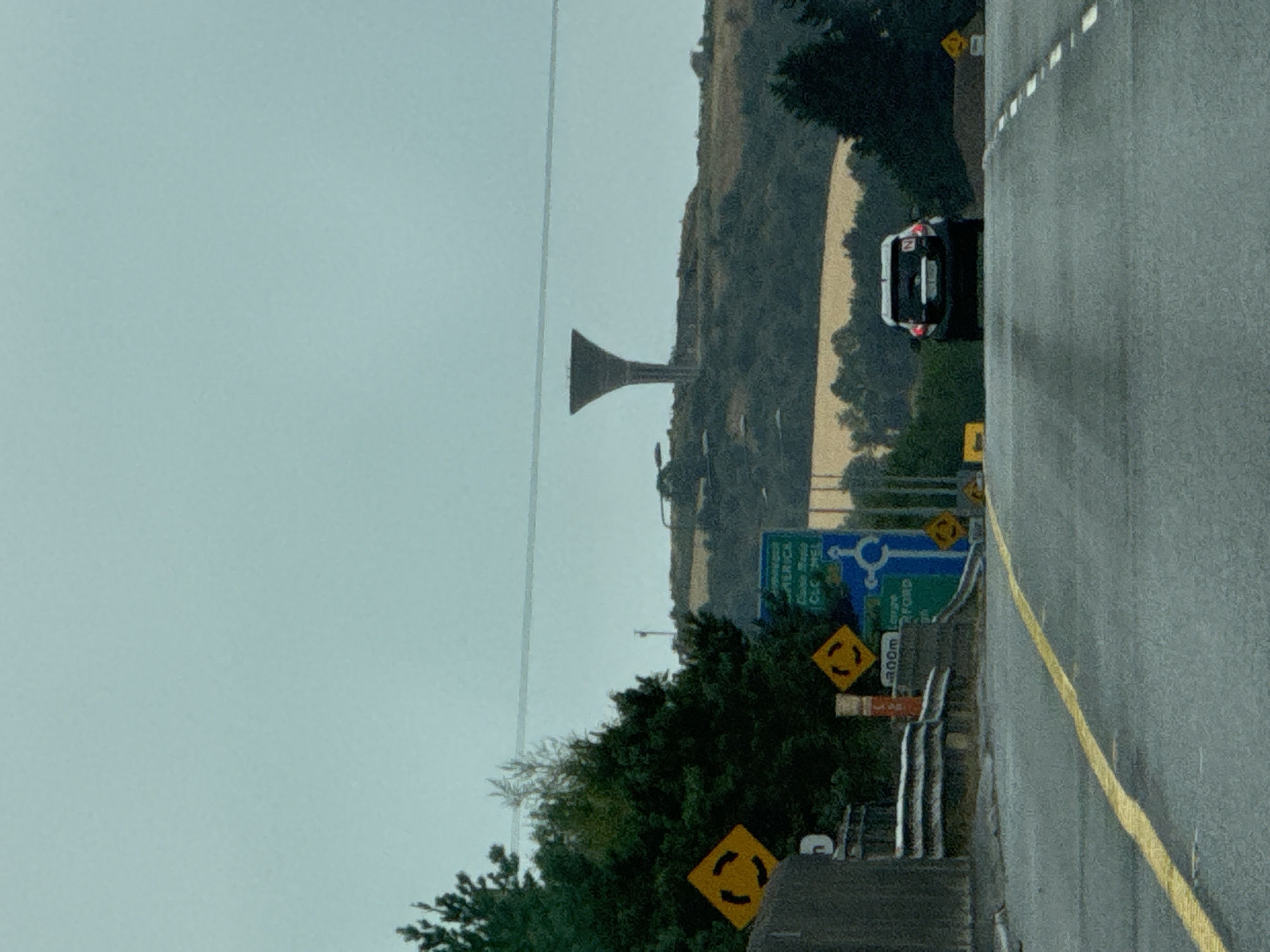 The image captures a view from a road in Kilmacow, Ireland. In the distance, a tall, conical water tower stands on a grassy hill. The sky is overcast, and the landscape is characterized by rolling hills covered in trees and fields. A dark-colored car is visible on the road ahead, approaching the camera. Road signs indicating a roundabout are present on the left side of the road, along with a larger blue directional sign pointing towards Limerick, Clara, and Kilkenny. The road itself is asphalt with a yellow line marking the center. The overall scene suggests a rural or semi-rural setting in Ireland.