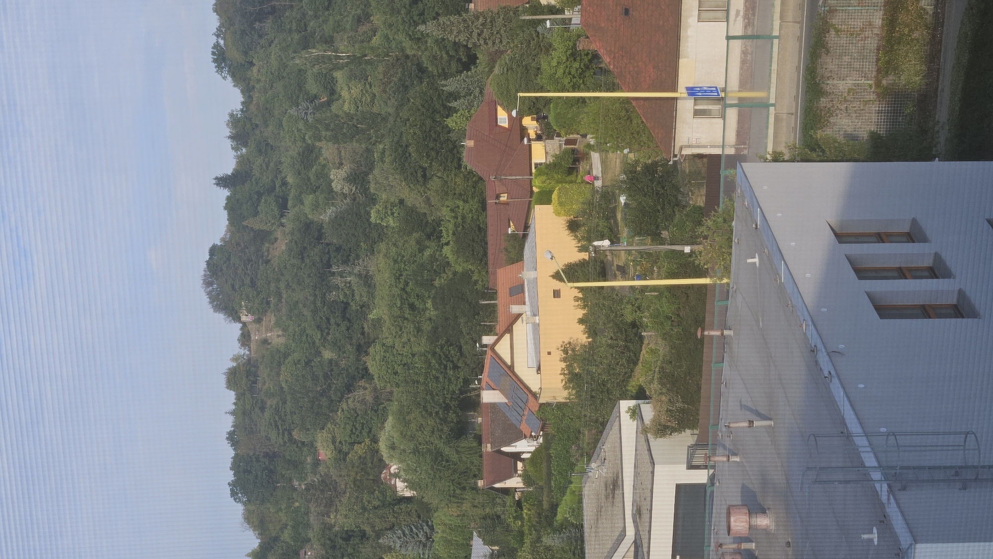The image captures an aerial view of a residential area with lush green trees and a hilly landscape in the background. In the foreground, a modern white building with multiple windows is visible. Adjacent to it, a grey roof with various protrusions and a metal ladder extends. Beyond these, several houses with red-tiled roofs and some with solar panels are nestled among the trees. A yellow pole stands prominently in the middle ground. Further back, a person in a pink top is walking near a yellow pole and some greenery. The sky is clear with a pale blue hue. The scene appears to be set in a suburban environment with a blend of residential buildings and natural foliage. The image is taken from an elevated perspective, suggesting it was captured from a higher building or a drone. The overall impression is one of a peaceful, daylit residential neighborhood. This image is of the area located near Fintice, a part of Prešov, Slovakia.