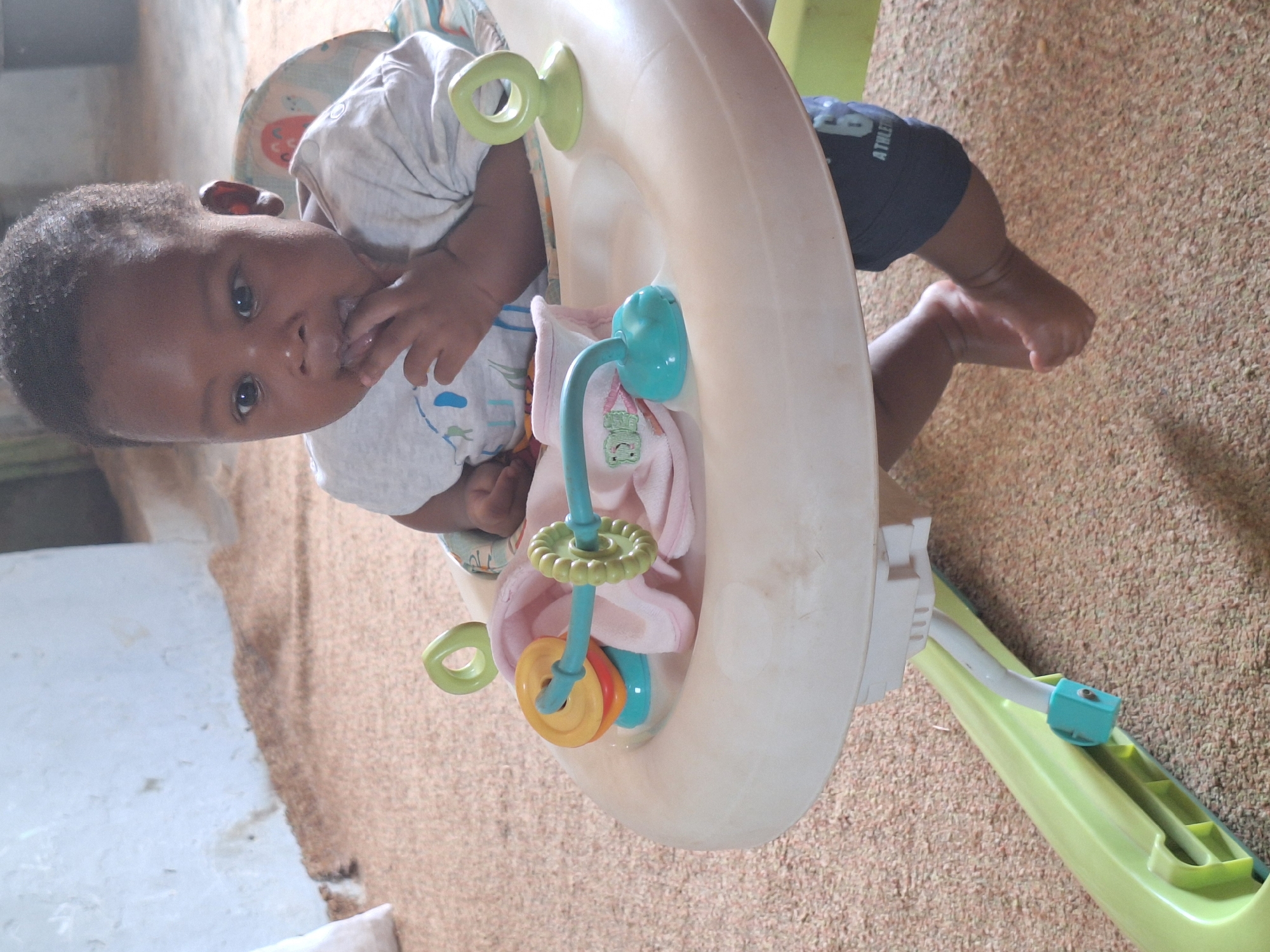 A baby sits in a walker with hanging toys in Sabon Tasha, Nigeria. The walker is ivory colored with green and blue accents. The baby is wearing a light-colored shirt and dark blue shorts. The floor is covered in a tan, textured material.