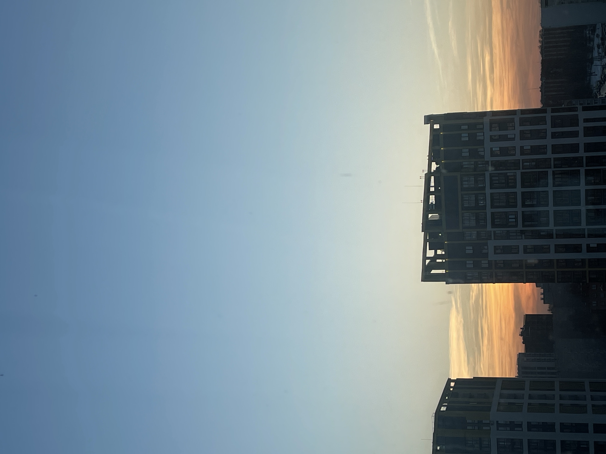 The image displays an urban residential landscape within Izhevsk, Russia, at either sunrise or sunset. The sky, occupying the upper two-thirds of the frame, transitions from clear blue overhead to a gradient of light blue, yellow, orange, and red near the horizon, where sparse, wispy clouds are present. Two prominent, modern multi-story buildings, consistent with residential apartment blocks, are visible in the lower portion of the scene. The building on the left is partially framed, showing a light-colored facade with darker recessed sections and numerous rectangular windows. The building on the right is more centrally positioned, featuring a similar architectural style with a light main structure, dark window sections, and complex, possibly decorative or unfinished, structural elements at its top. In the mid-ground, between and behind these two structures, darker silhouettes of additional urban buildings and indistinct vegetation are visible against the illuminated horizon. No people, specific actions, or interactions are observable in the scene.