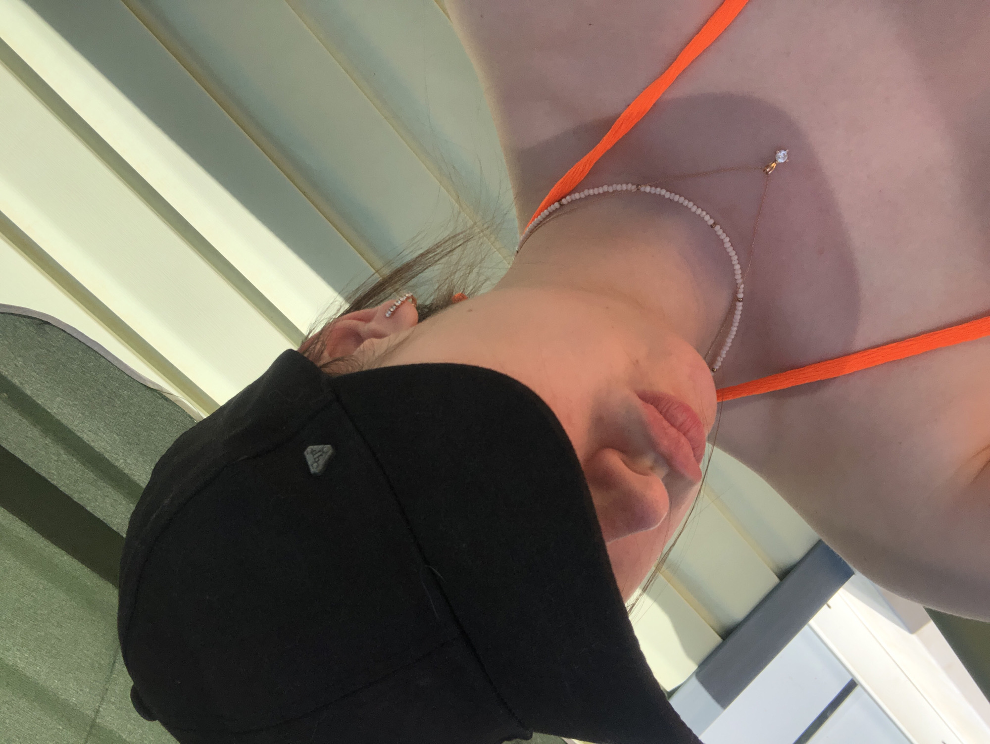 A woman wearing a black baseball cap and orange bikini top is pictured from a low, angled perspective. She also wears a delicate necklace and earrings. The background appears to be a structure with white and green slats. The location is unknown, and cannot be determined with the data provided.