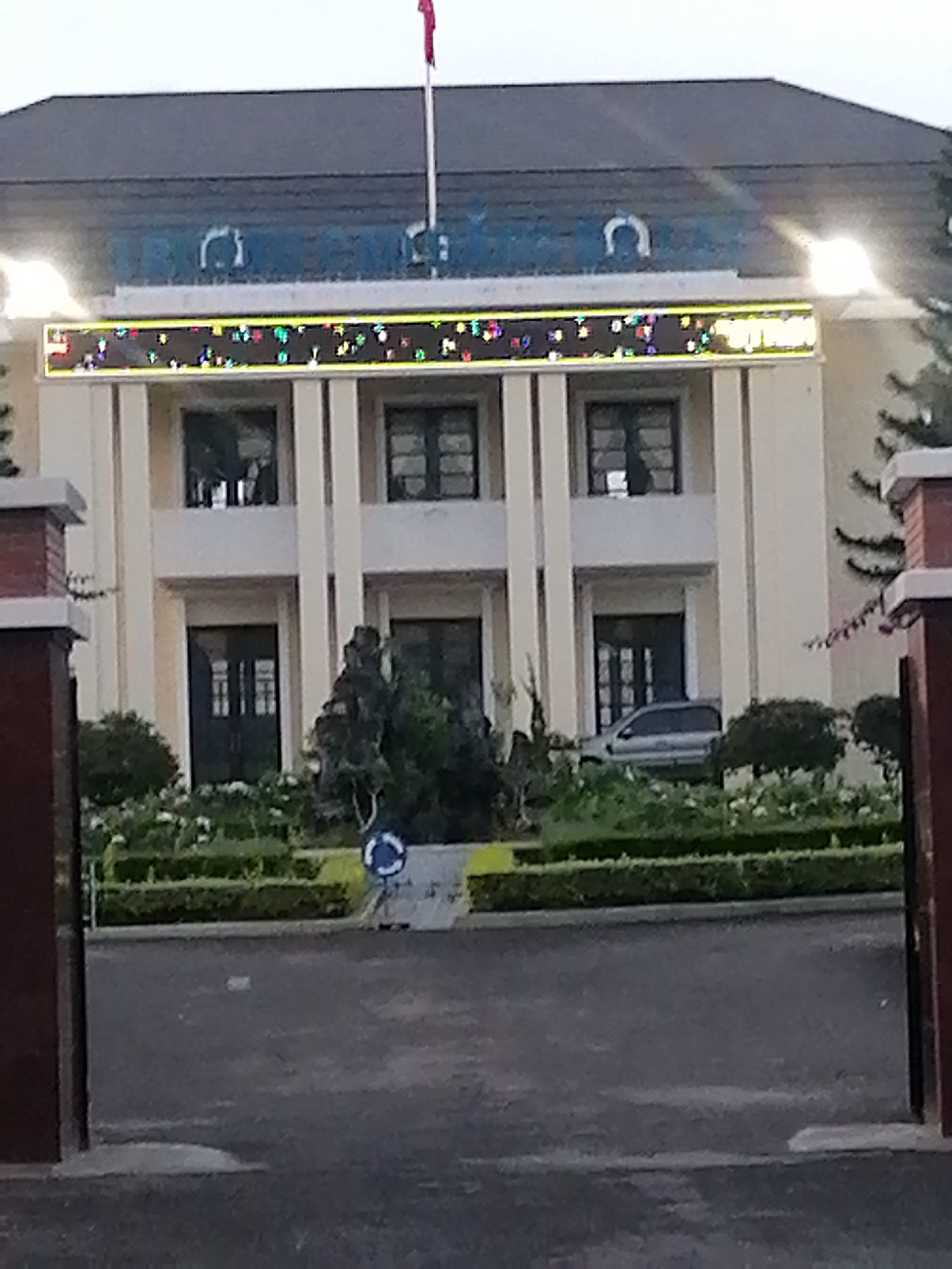 The image shows the exterior of a building, likely an administrative or institutional building, in Da Lat, Vietnam. The building is a pale yellow color with a dark gray roof and features a prominent LED display sign above the ground floor windows. The sign displays colorful, abstract patterns, suggesting it might be used for announcements or decorative purposes. There are several large windows and doors on the facade. In front of the building, there is a manicured garden with trimmed hedges and flowering plants. A portion of a silver car is visible parked to the right of the building entrance. A circular blue and white traffic sign is situated in the garden area. The overall scene appears to be a well-maintained public space.