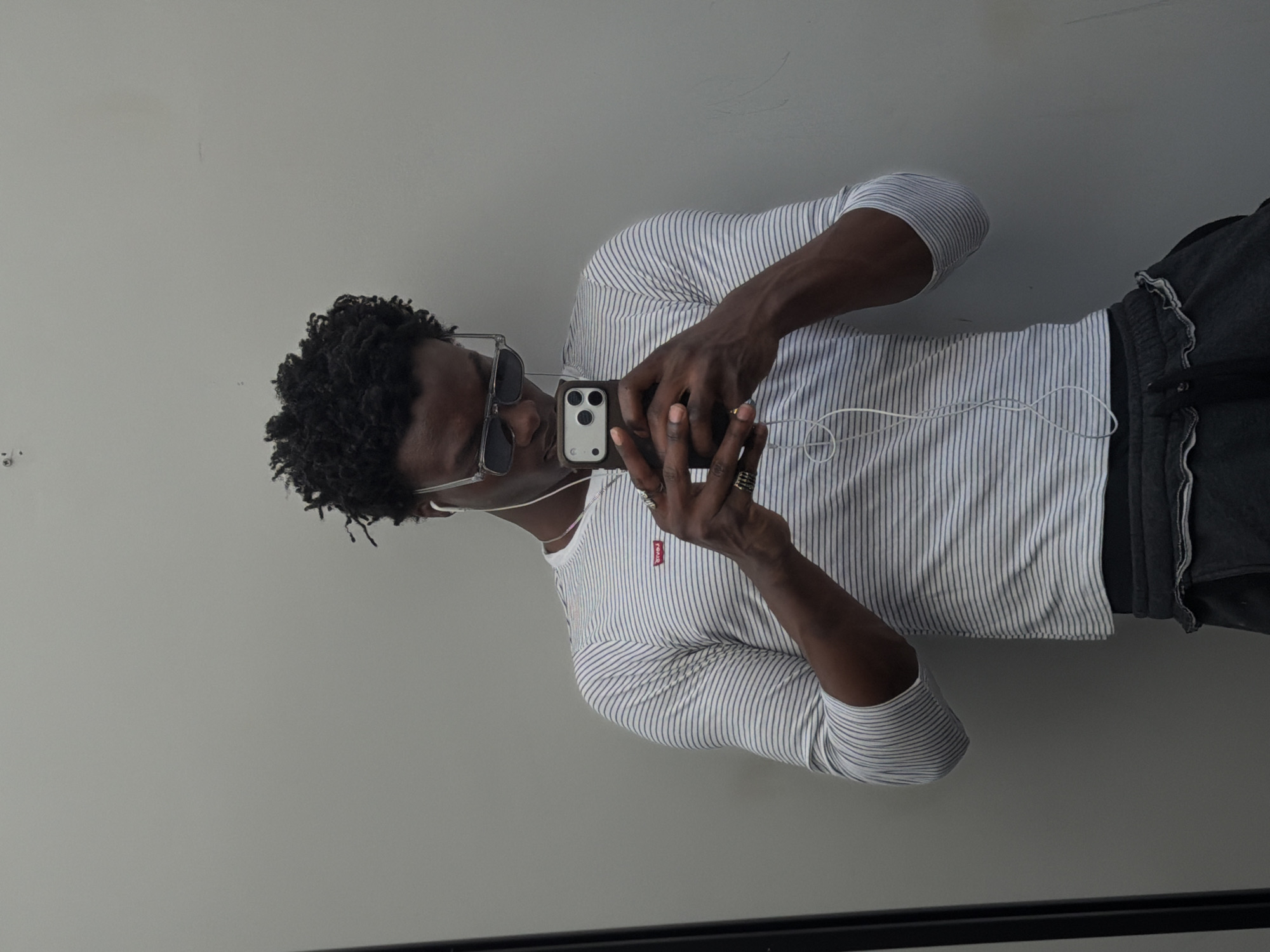 A man with dark curly hair and sunglasses is taking a selfie in a room with a gray wall. He's wearing a striped long-sleeved shirt with a small red tag on the chest and dark-colored shorts with ruffled edges. He is holding a phone with both hands. Part of a black frame is visible at the bottom of the image. There is no indication of the image being taken in London or the United Kingdom.