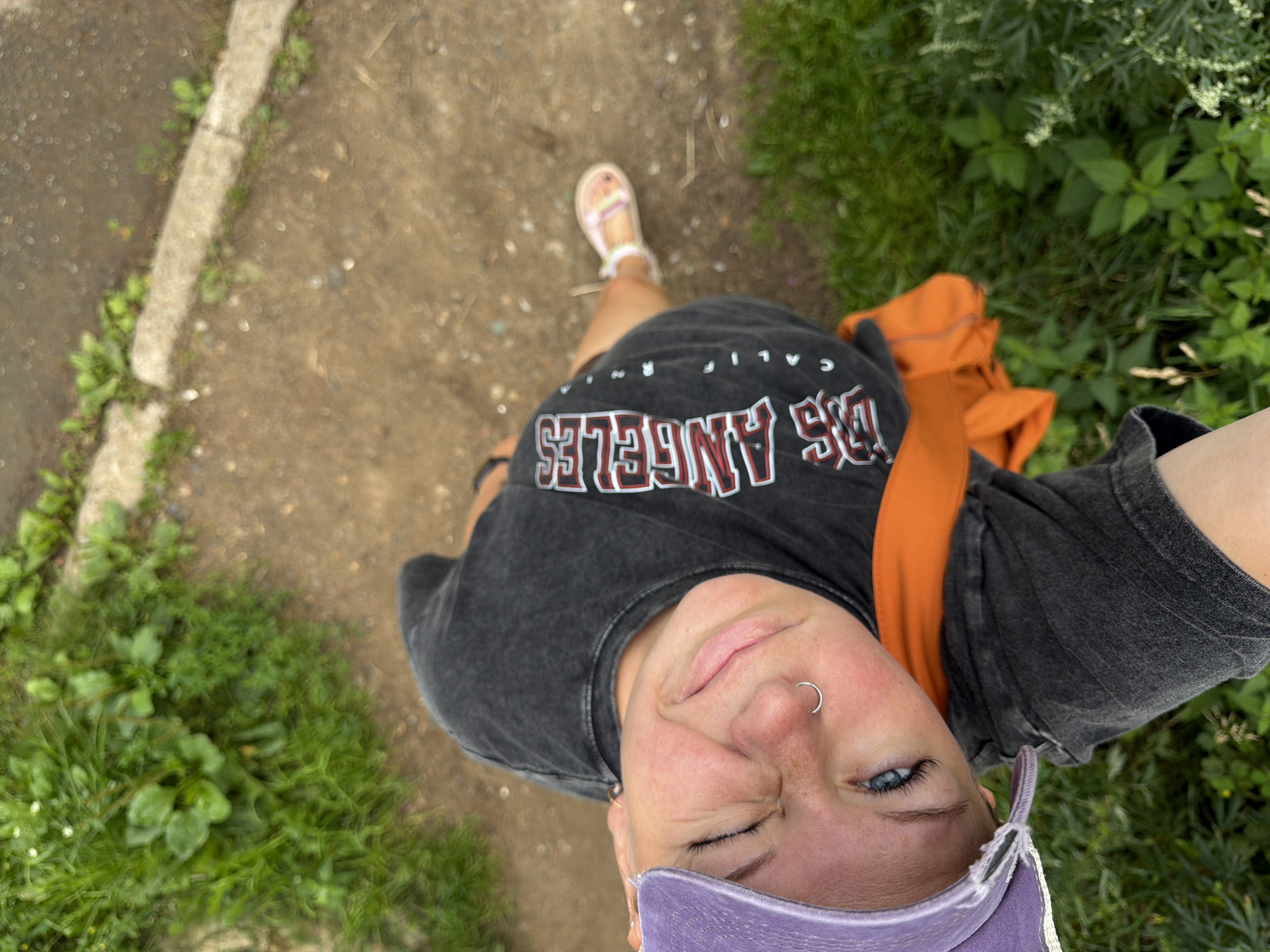 A selfie taken from above shows a young woman outdoors, looking up at the camera with her eyes closed and a slight smile. She is wearing a grey t-shirt with "LOS ANGELES" printed on it in red and white lettering, a purple baseball cap, and an orange garment draped around her shoulders. A silver nose ring is visible on her left nostril. Her right foot, clad in a sandal, is positioned forward. The background consists of a dirt path with some greenery and a concrete edge. The image does not provide any specific visual cues to determine its location within the city of Chaikovsky, Russia.