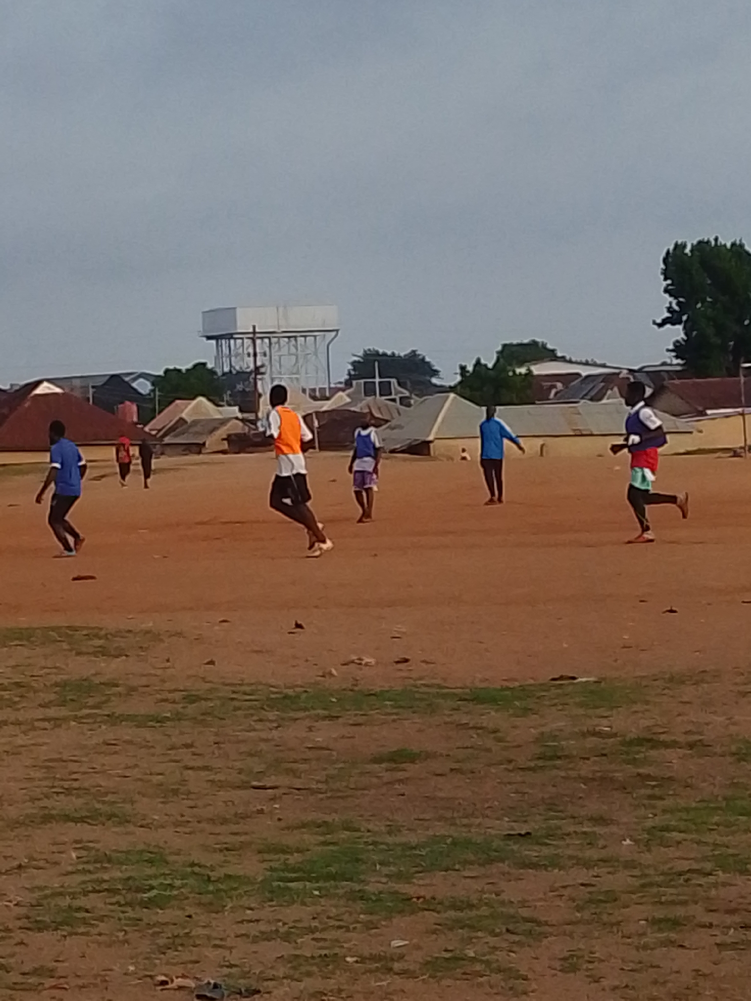 The scene is an outdoor setting, likely a field or dirt lot in Bukuru, Nigeria, where a group of individuals is engaged in what appears to be a casual soccer or football game. There are seven people visible in the image, wearing different colored shirts. One person is wearing an orange vest, others are in blue and white jerseys. All of them are dark-skinned. They are distributed across the field, with some running. The ground is primarily dirt with some scattered patches of grass. In the background, there are low buildings with corrugated metal roofs, a water tower, and some trees. The sky is overcast.