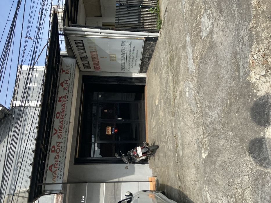 The image shows the exterior of a law firm in Purwakarta, Indonesia, located at the coordinates -6.555 latitude and 107.437 longitude. The firm is called "WARISON SIMARMATA, S.H., M.H." and is located at Jln. Jend. A. Yani No. 124/65. The sign above the entrance reads "ADVOKAT" and the sign to the right reads "KANTOR ADVOKAT/PENGACARA". A black motorcycle is parked in front of the entrance, with a sign in Indonesian that reads "BUKA" (open). The entrance has two glass doors with black frames. There is a partial view of a white car parked to the left of the entrance. The building is surrounded by a concrete sidewalk with a small patch of grass visible on the right side of the image. There are multiple electrical wires running across the top of the image. The environment is urban, with buildings and sidewalks visible in the background.