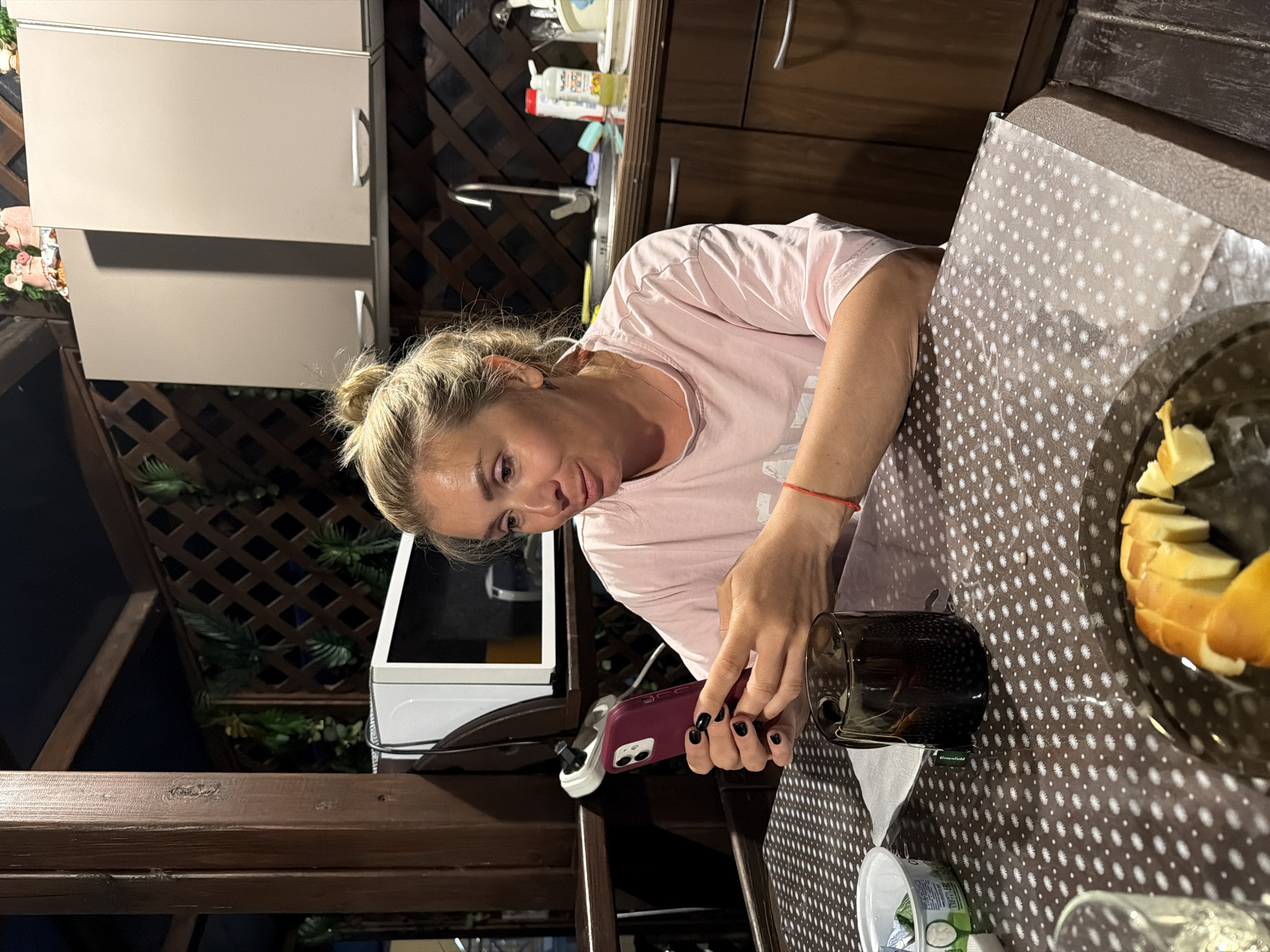 In a covered outdoor kitchen area that appears to be part of a private residence in Sochi, Russia, a blonde woman with her hair tied up in a bun is seated at a table. She is wearing a pink t-shirt and looking at her smartphone, which she holds in her hands. Her nails are painted black. On the table in front of her is a dark glass mug with a tea bag, a small white container, and a plate of sliced pastry or bread. In the background, there are kitchen cabinets, a microwave oven, and decorative greenery. The overall impression is of a casual, domestic setting.