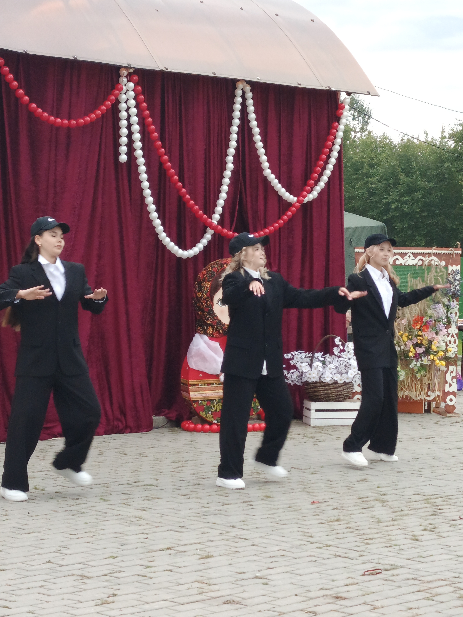 Three young females in matching black suits and white shirts, each wearing a black baseball cap, are performing a synchronized dance on a paved outdoor area. They are positioned in front of a stage draped with dark red curtains and adorned with strings of red and white beads. To the right of the stage, a large, colorful, traditional Russian nesting doll is visible. A woven basket filled with white flowers sits on a white crate in the middle ground. In the background, green trees and a cloudy sky are visible, suggesting an outdoor event. The event appears to be taking place in or near the village of Medvedyovka, Russia, possibly during a festival or cultural celebration, given the traditional elements present.
