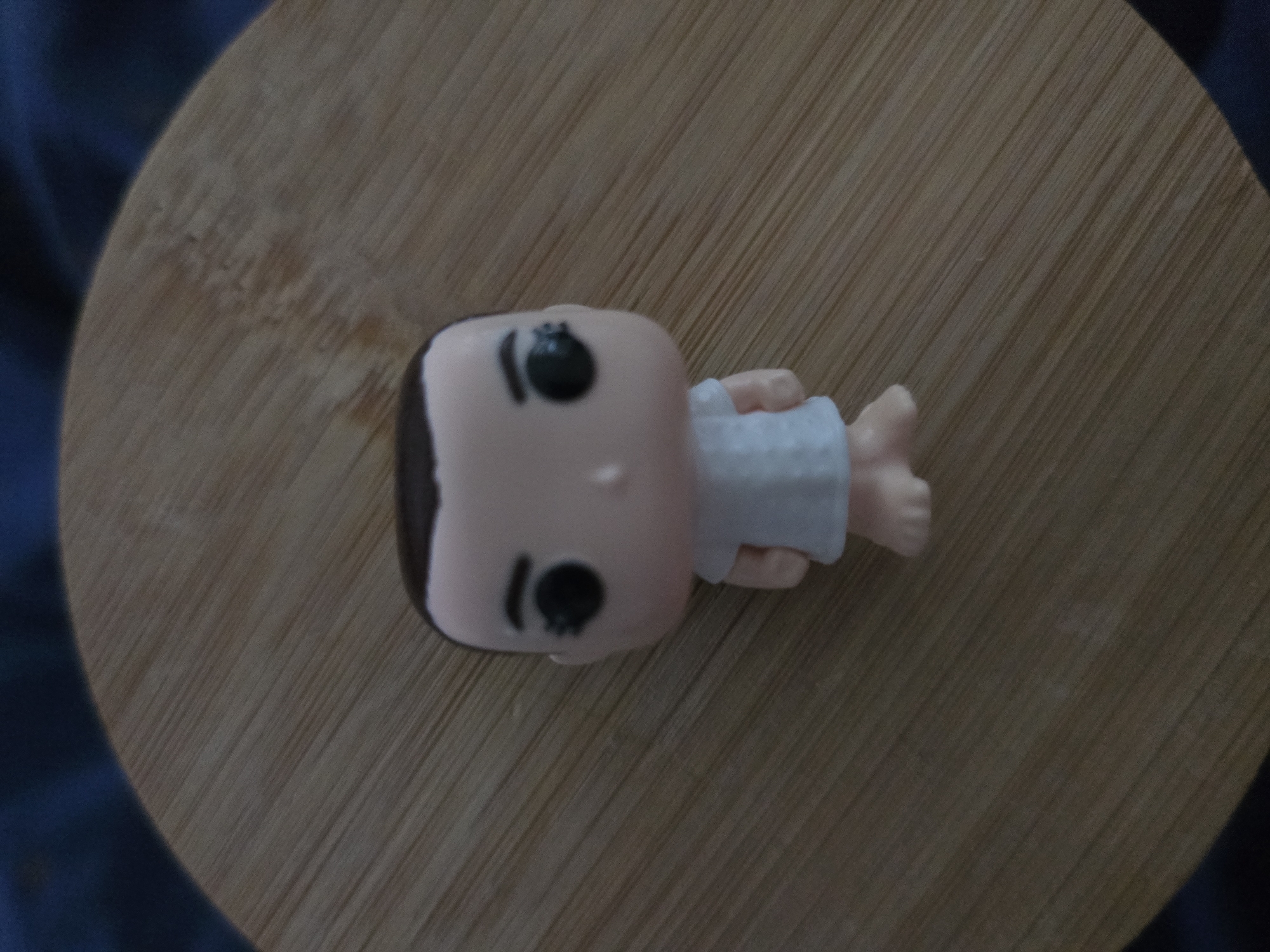 A Funko Pop doll, resembling the character Eleven from Stranger Things, is positioned centrally on a circular wooden surface. The doll has short black hair, large black eyes, and is wearing a simple white dress. Its hands are held slightly away from its body, and its legs are together. The wooden surface has a visible grain pattern and appears to be a placemat or a decorative coaster. The background is out of focus and appears to be a dark blue fabric. There are no indications of the location being in Petropavlivska Borshchahivka, Ukraine.