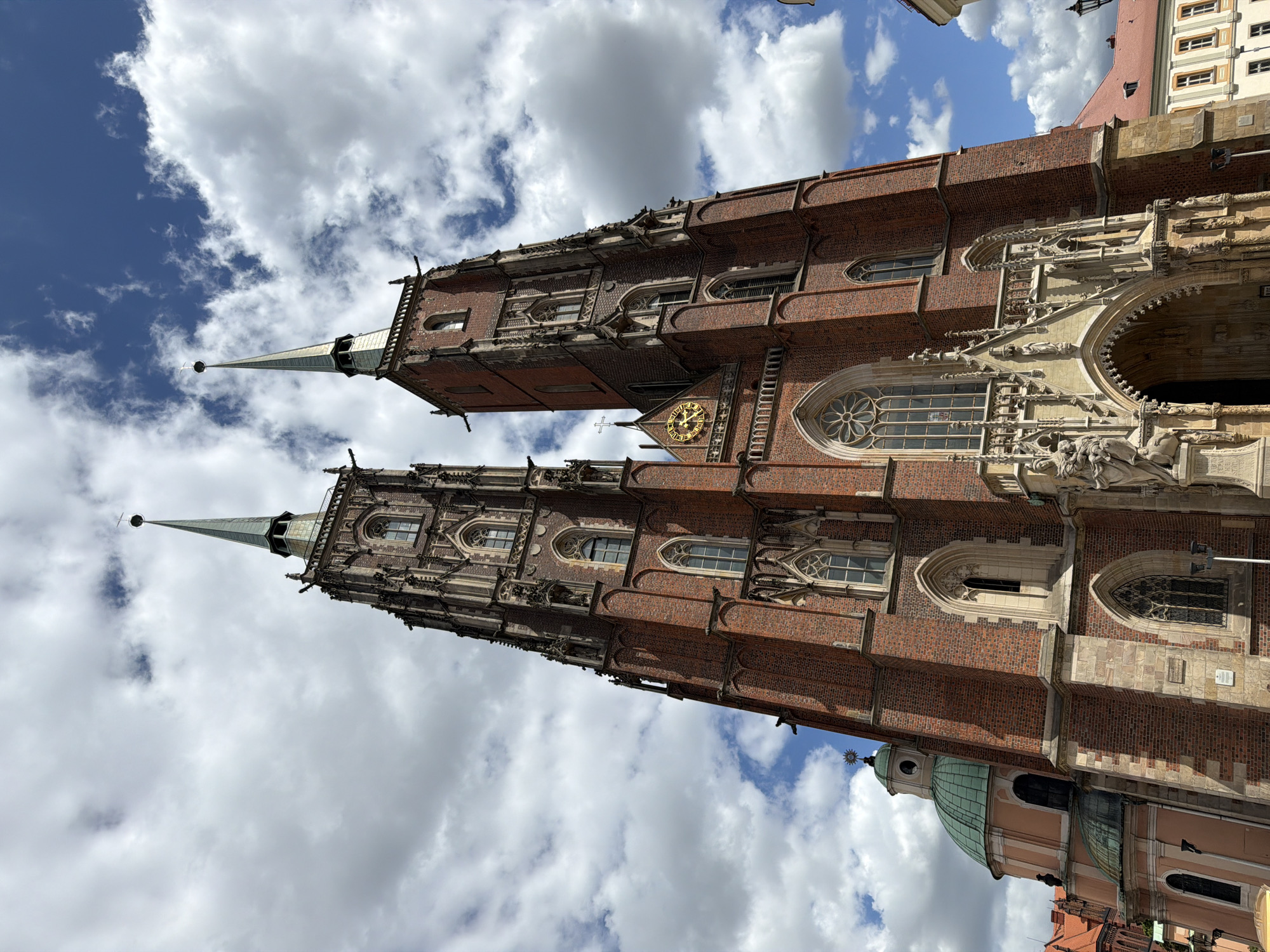 A low-angle shot captures the twin spires of a large, Gothic brick cathedral reaching towards a partly cloudy blue sky. The cathedral's facade features intricate stone carvings around a large arched entryway, a circular rose window above it, and numerous gothic-style windows. A gold clock face is visible high on one of the towers. To the left, a smaller green-domed building is partially visible. The image appears to be taken from a public square, with hints of surrounding buildings and a street lamp in the lower right corner. The overall scene suggests a historically significant architectural landmark.
