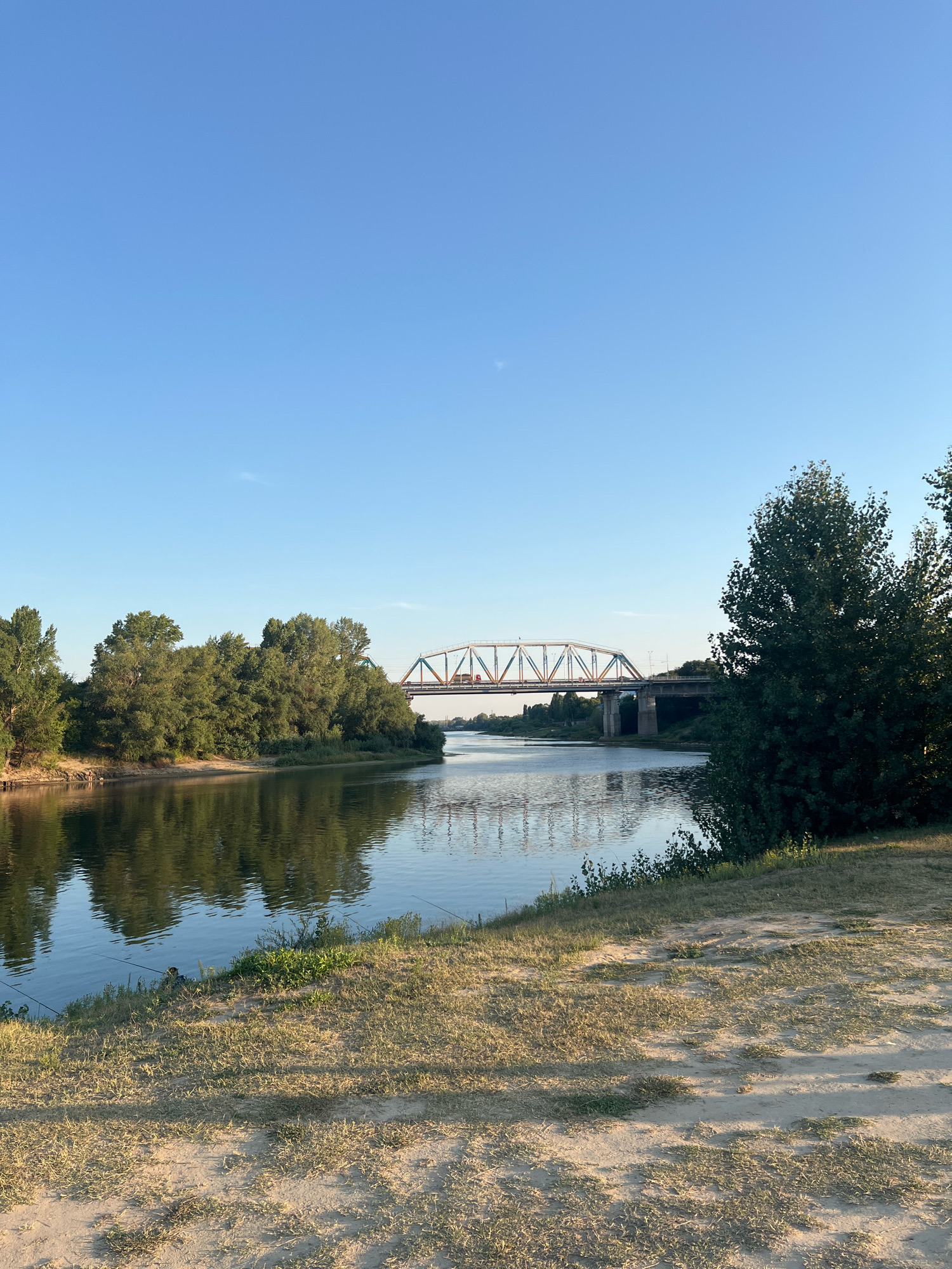 The image shows a wide river flowing under a bridge in Bender, Moldova. The sky is clear and blue. Trees and vegetation line the riverbanks. The water reflects the trees and the bridge. There is a dirt path in the foreground with dry grass. The bridge appears to be a railway bridge or a road bridge with metal trusses.