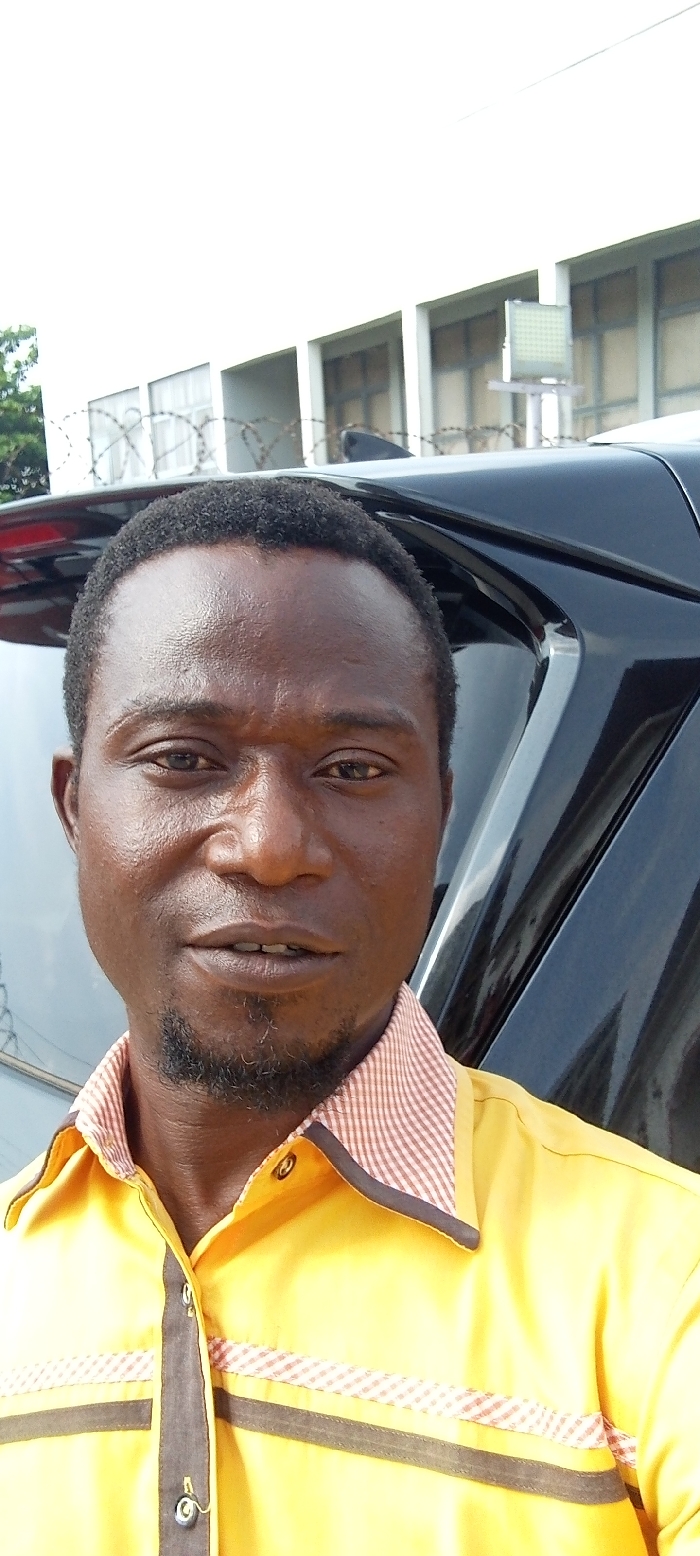 A dark-skinned adult male is depicted in a close-up, chest-up portrait, likely a selfie, captured in Akure, Nigeria. The male has short, dark, curly hair and a short, dark goatee. He wears a yellow collared shirt featuring a red and white checkered pattern on the inner collar and two horizontal bands across the chest: an upper checkered band and a lower dark brown/black band. His expression is neutral, with his eyes directed towards the camera.
In the background, to the subject's left, a portion of a black vehicle is visible, including part of its roofline and rear window. Behind the vehicle and extending across the frame is a multi-story white building with multiple rectangular windows. Barbed wire runs horizontally in front of the building's lower windows, indicating a secure perimeter. A large, square-shaped floodlight or security light, composed of a grid of smaller lights, is mounted on a pole above the barbed wire and near the building's roofline. Green foliage is partially visible on the far left of the background. The scene appears to be outdoors during daylight hours.