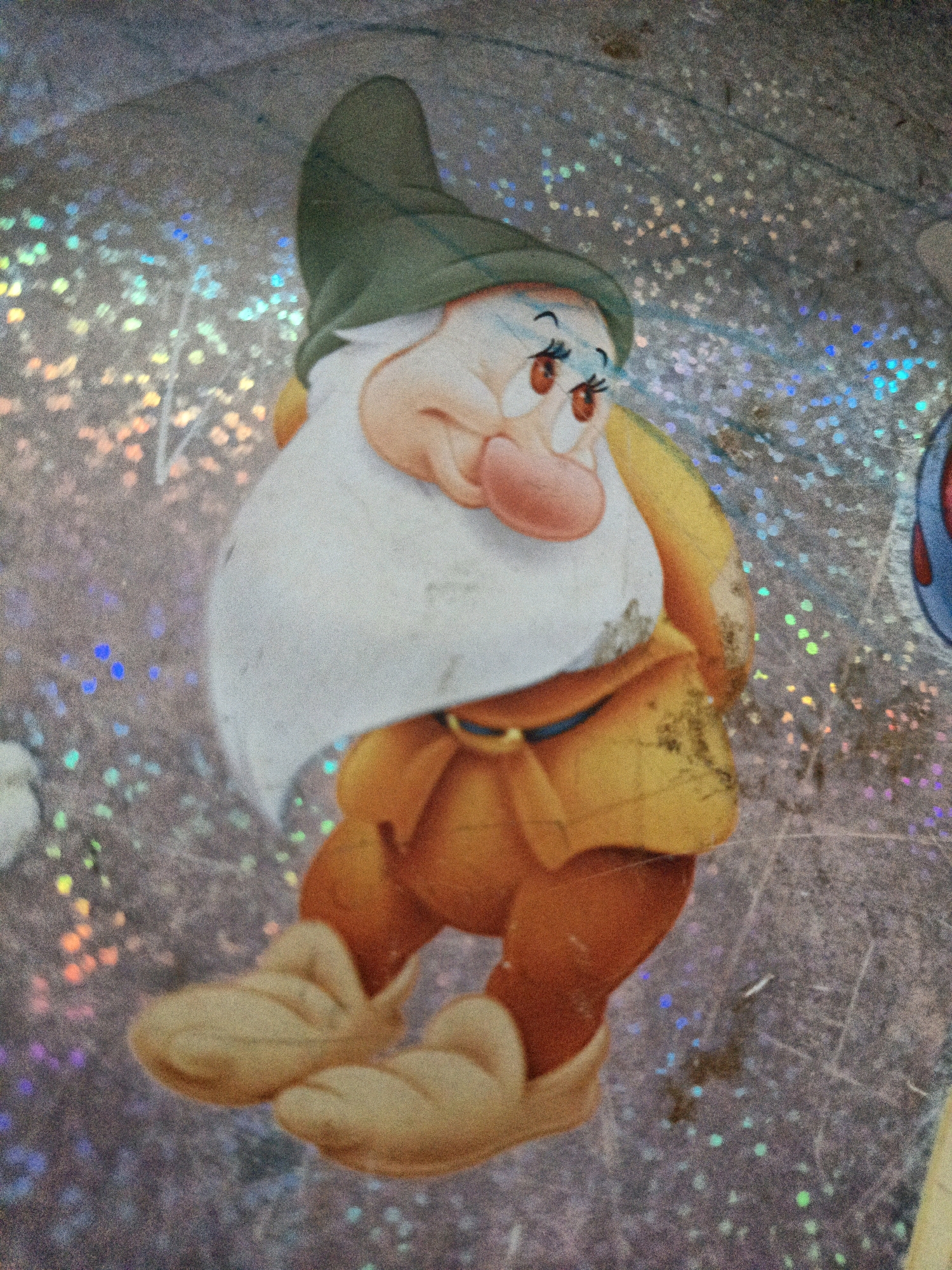 The image shows a close-up of a sticker featuring a cartoon character, likely one of the Seven Dwarfs from Disney's Snow White. The character is depicted with a green hat, a large white beard, rosy cheeks, and is wearing a yellow tunic over orange pants and brown shoes. The sticker appears to be applied to a textured, holographic surface that shimmers with colorful, fragmented light. There is some wear and tear visible on the sticker, with scuffs and dark marks present. The background is a blurred pattern of iridescent specks of light. No specific location in West Jakarta, Indonesia, can be determined from this image.