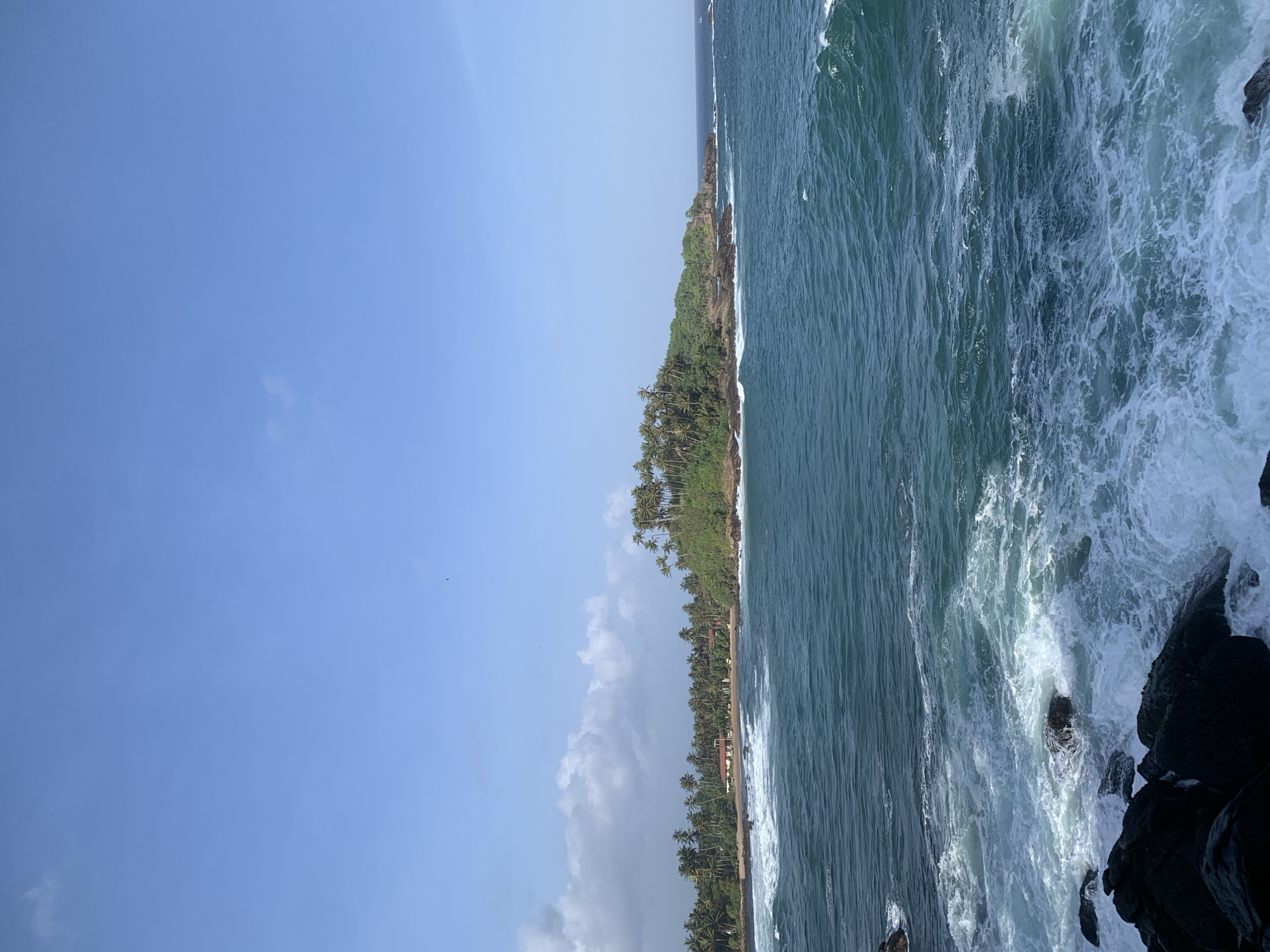 This is a landscape photograph taken in Dondra, Sri Lanka, featuring a body of water, a coastline with vegetation, and a clear sky. In the foreground, dark, rugged rocks are partially submerged in the ocean, with waves crashing against them and creating white foam. The water is a deep blue-green, with visible ripples and currents.
The midground shows a curved coastline dotted with numerous palm trees and other lush green vegetation. A small beach area is visible, with a few buildings featuring red-tiled roofs nestled among the trees. The vegetation covers a hilly terrain that extends into the sea on the right side of the image.
The background consists of a vast, pale blue sky with a few scattered white clouds, primarily on the left side. The overall scene depicts a tropical coastal environment.