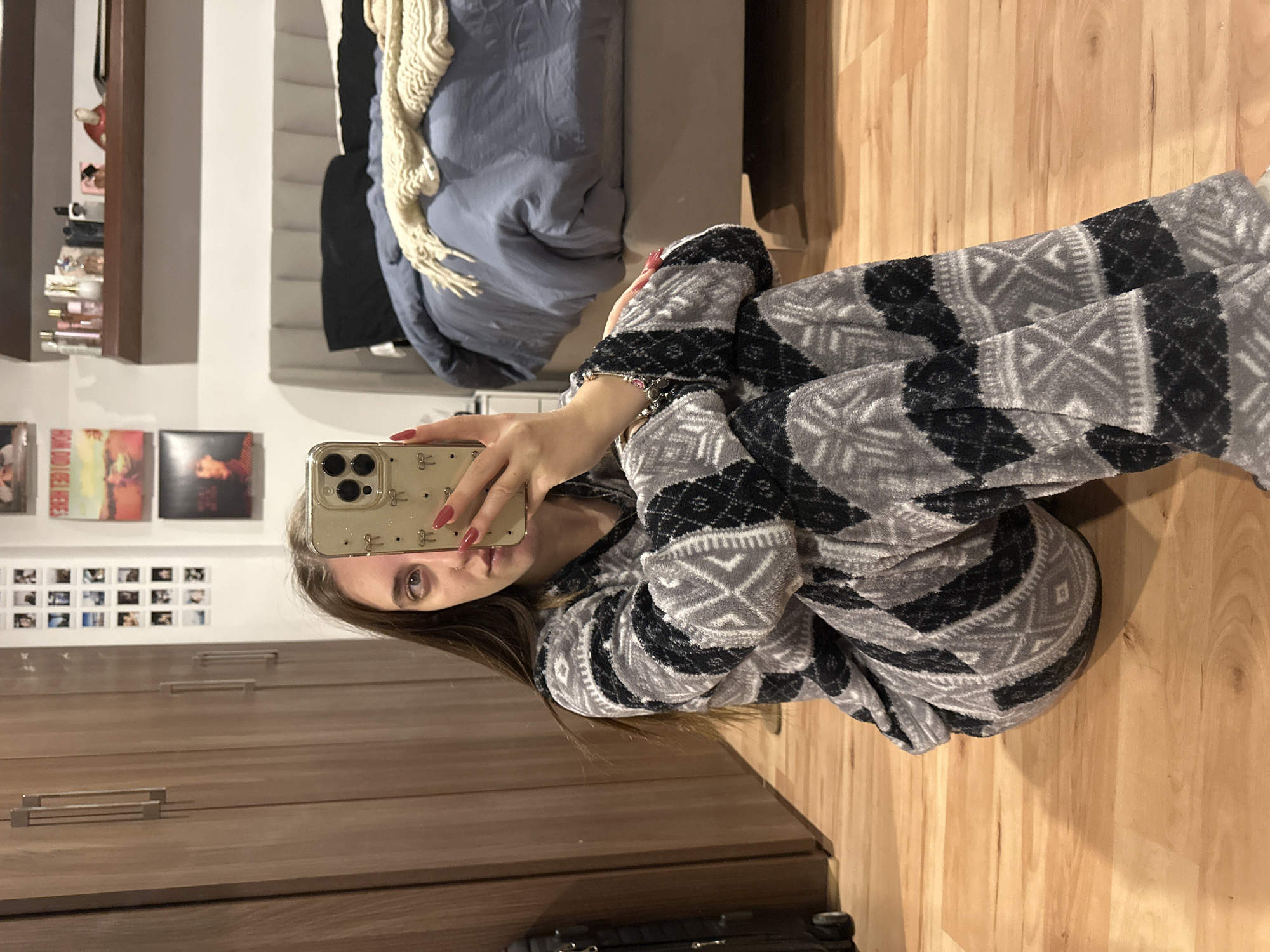 A young woman is taking a selfie indoors. She is wearing patterned grey and black pajamas. The background includes a bed with a blue duvet, a wooden floor, a closet with metal handles, and a wall with a shelf, album covers, and polaroid photos. The location is unspecified in Huixquilucan de Degollado, Mexico, but the scene is likely a residential bedroom.