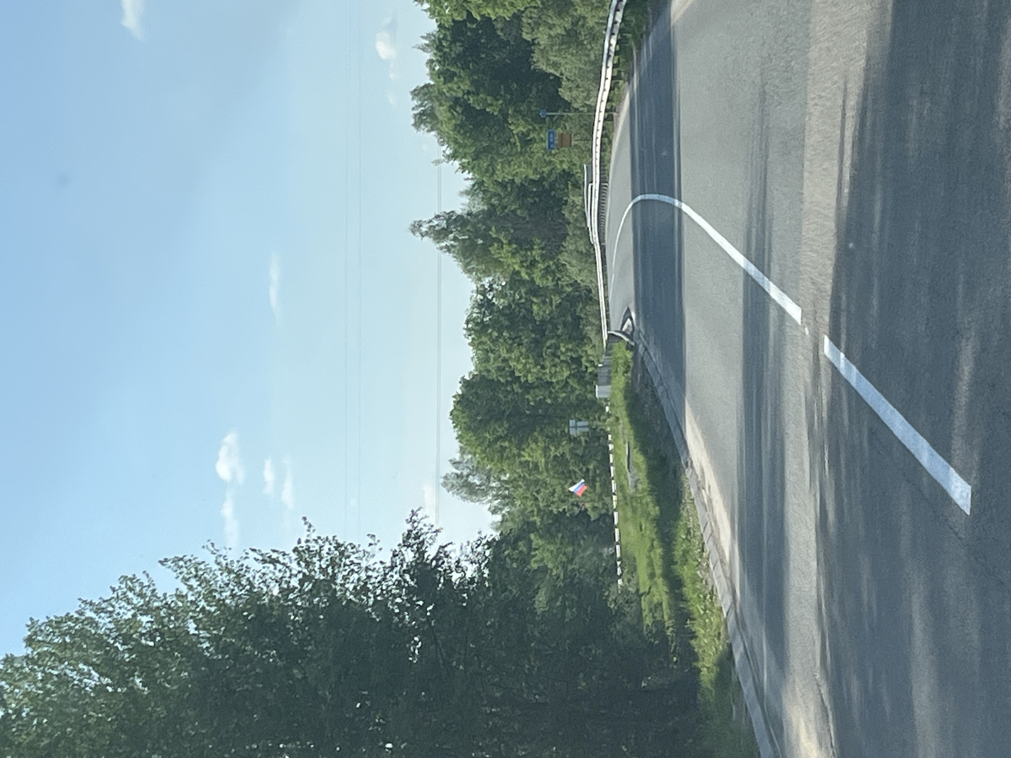 The image shows a roadside scene in Izhevsk, Russia. An asphalt road curves to the right, marked with a solid white line. A metal guardrail runs along the road's edge. A grassy area and trees border the road. A Russian flag is visible in the background, mounted near a post. The sky is light blue with some clouds. Overhead power lines run parallel to the road.