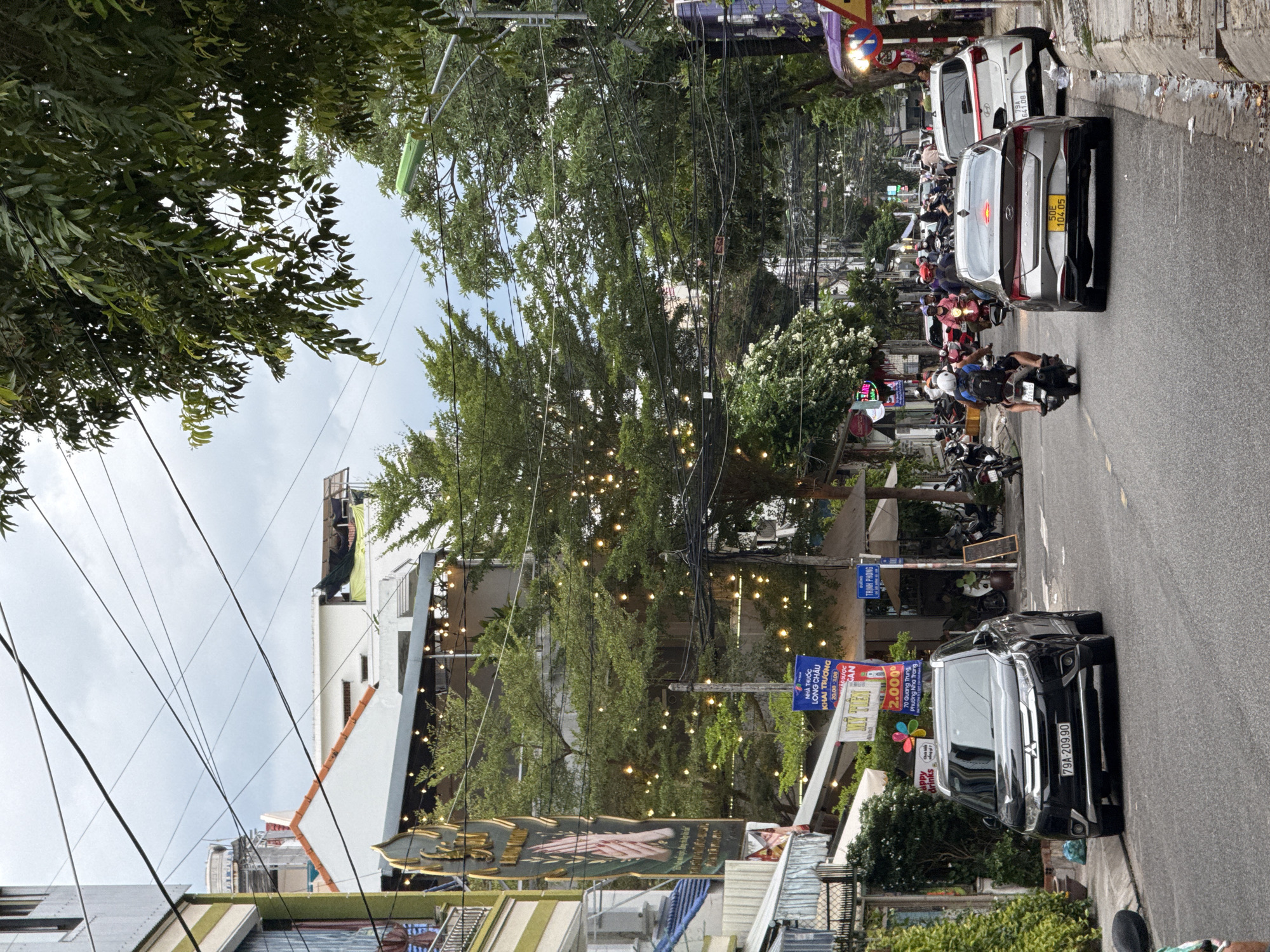 The image depicts a street scene in Nha Trang, Vietnam, characterized by a mix of urban elements and lush greenery. The street is paved, with a raised concrete curb along the right side. Overhead, a dense network of electrical wires crisscrosses the sky, a common sight in Vietnamese cities.
On the left side of the street, several buildings are visible. One building has a distinctive green and gold sign with Vietnamese text and an image of hands. Other structures appear to be shops or restaurants, indicated by awnings and signage. String lights are draped across the facades, suggesting a festive or commercial atmosphere. A black Mitsubishi Outlander SUV is parked parallel to the curb on the left.
The right side of the street features a row of buildings and trees. A white Hyundai hatchback is parked on the right side of the road. A traffic light pole with a green fixture is visible further down the street, along with various signs, some with Vietnamese text and warning symbols.
The street is active with traffic. Several motorcycles are present, with riders in various positions. One motorcycle is being ridden by a person wearing a helmet and a blue backpack. Other vehicles are seen in the background, indicating moderate traffic flow. Trees with dense green foliage line the street and overhang the buildings, providing shade and a touch of nature to the urban landscape. The sky is overcast, suggesting a cloudy day. The overall impression is a vibrant, bustling street scene in a Vietnamese city, blending modern urban life with natural elements.