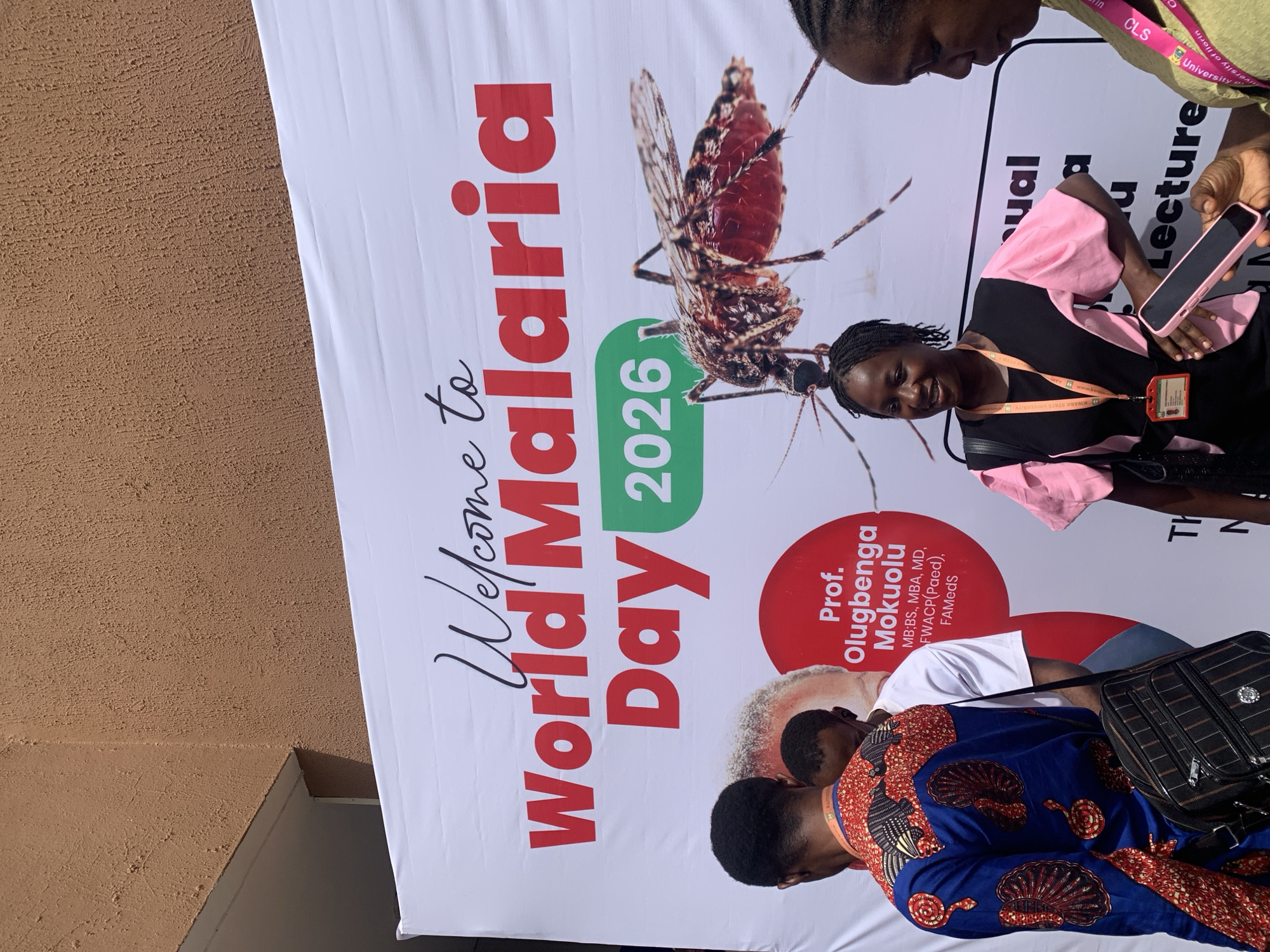 An outdoor scene captured with a slightly rotated perspective shows several individuals gathered around a large banner advertising "World Malaria Day 2026." The banner, predominantly white with red and green text, features a prominent image of a mosquito.
In the foreground, two individuals are partially visible, one wearing a vibrant blue and orange patterned top and carrying a black checkered bag, and another with their back to the camera, wearing a white t-shirt. To the right, a woman in a black and pink top smiles faintly, looking slightly upwards. She wears an ID badge around her neck, which appears to have "KWARA STATE UNIVERSITY" visible. Another person, seen from the chest up, is on the far right, holding a pink-cased smartphone. A person in the upper right corner, wearing a light green top with a pink lanyard, is also partially visible, with "University of Ilorin" discernible on the lanyard.
The banner's text includes "Welcome to World Malaria Day 2026." A red circular element on the banner introduces "Prof. Olugbenga Mokuolu," followed by his credentials: "MB;BS., MBA, MD, FWACP(Paed), FAMedS." Other partial text on the banner includes "sual," "Lecture," "CLS," "University of Ilorin," and "TH N." The background beyond the banner is a textured, light brown wall. The lighting suggests it is daytime. The people appear to be either attending or arriving at an event related to World Malaria Day, possibly in Ilorin, Nigeria, given the mention of Kwara State University and the University of Ilorin.