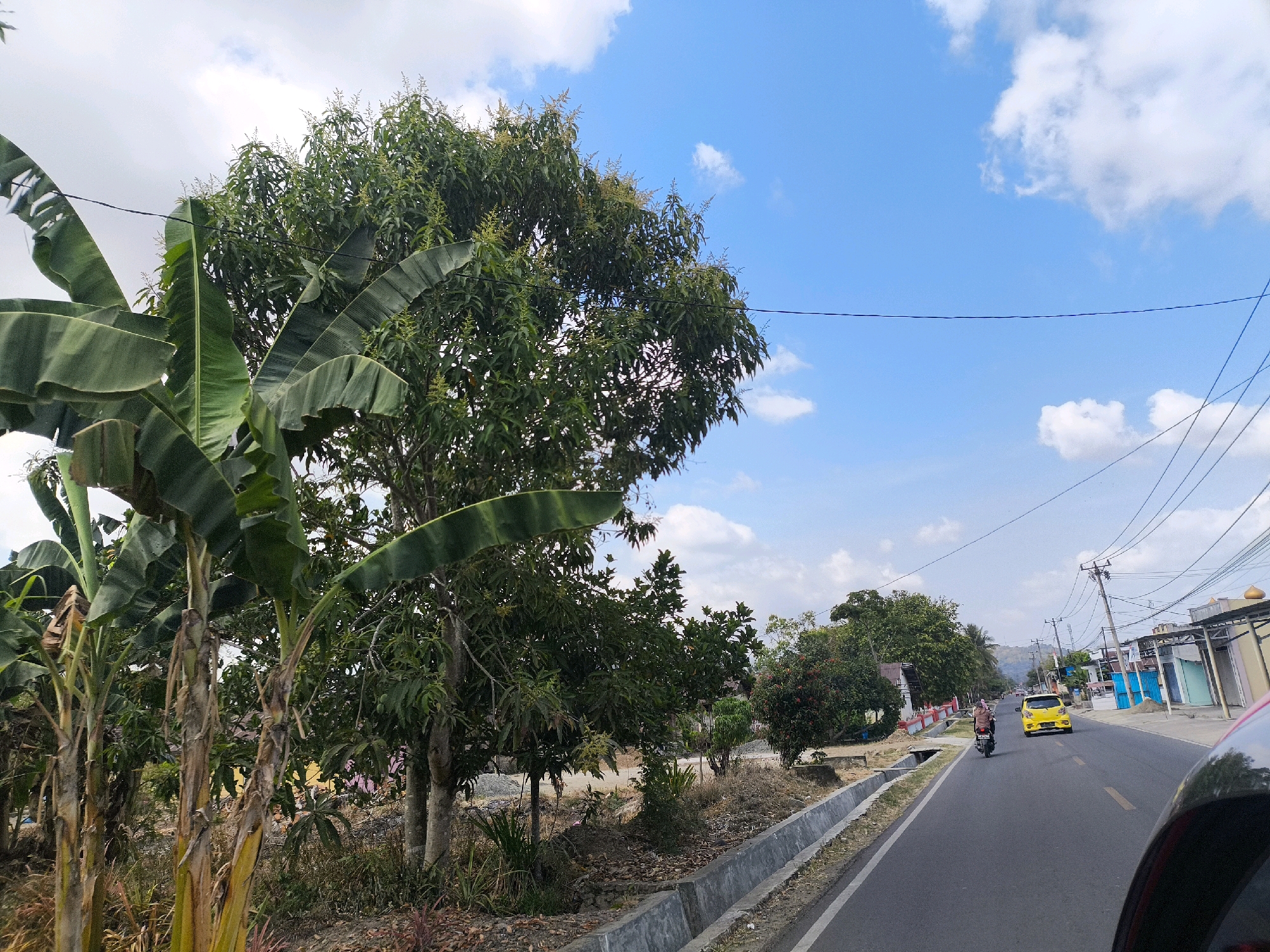 The image depicts a street scene in Talumopatu, Indonesia. A paved road runs from the foreground towards the horizon, lined on the left by a concrete drainage ditch and a variety of trees and vegetation, including prominent banana trees with large leaves and a leafy deciduous tree. On the right side of the road, there are buildings, including structures with colorful facades and a building with a golden dome. A yellow car is driving away from the viewer on the road, and a person on a motorcycle, also moving away, is in front of it. Overhead, power lines are strung between poles. The sky is blue with scattered white clouds. The scene appears to be set during daylight hours.