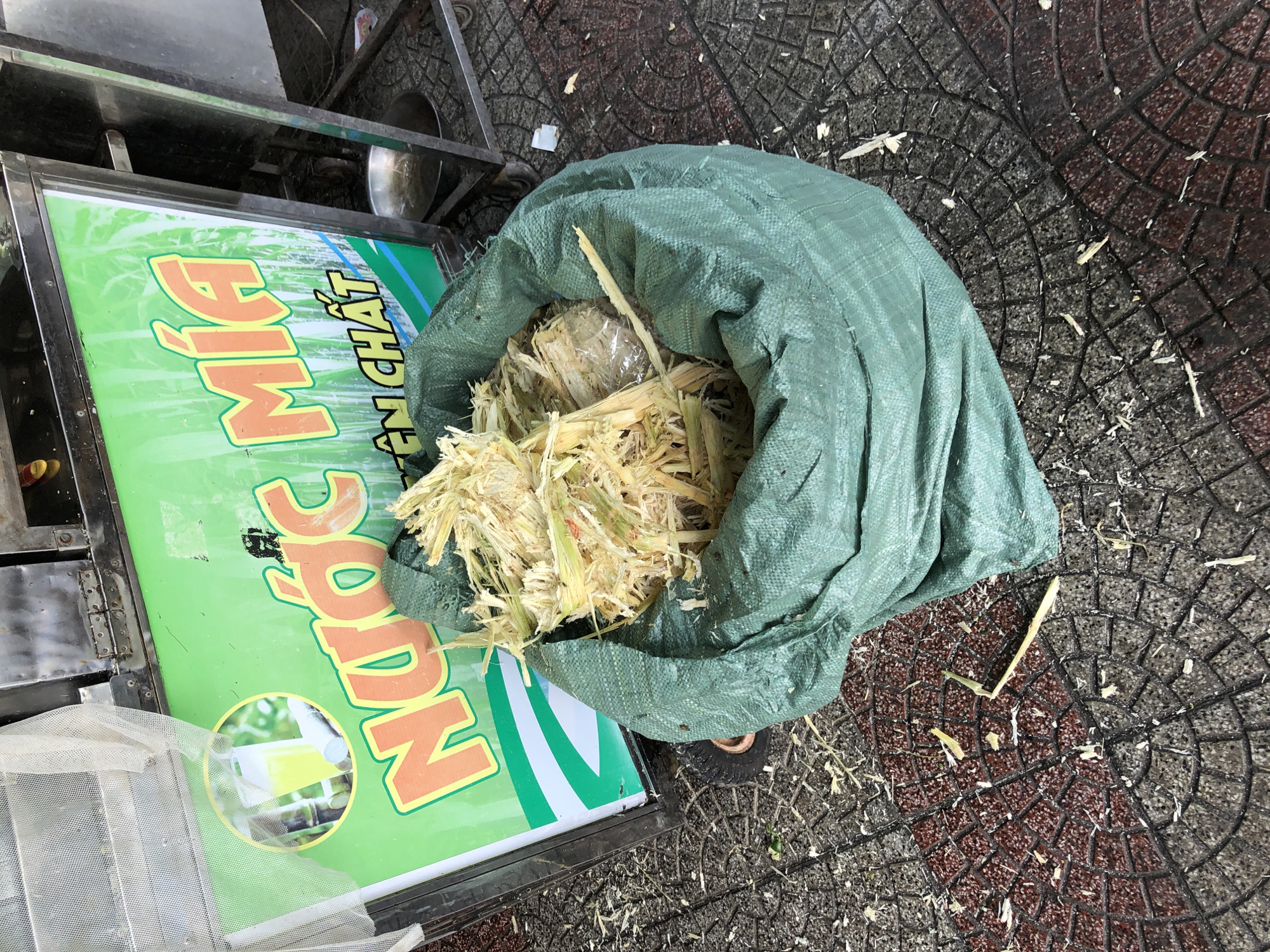 In the city of Đà Nẵng, Vietnam, an outdoor scene depicts a sugarcane juice stand. A green tarp bag filled with fibrous sugarcane pulp sits on a tiled sidewalk. Behind the bag is a sugarcane juice machine with a green sign that reads "NƯỚC MÍA" in large orange letters, meaning sugarcane juice. The sign also features an image of a glass of sugarcane juice. The sidewalk is made of interlocking tiles, some of which are red and others gray. Scattered on the ground are remnants of sugarcane pulp. The scene appears to be captured during daylight.