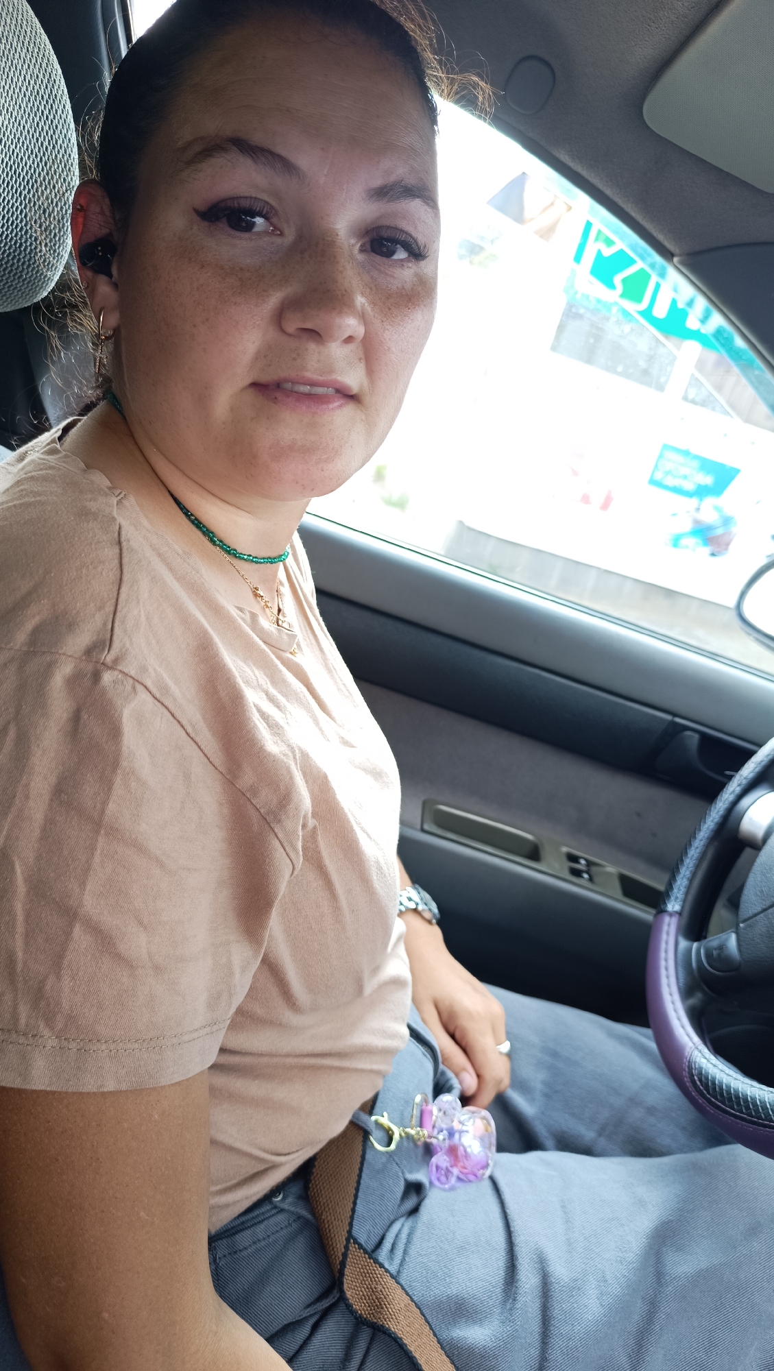 A woman is seated in the driver's seat of a car. She is wearing a tan t-shirt, grey jeans, and a watch on her left wrist. She has a necklace with green beads and a gold pendant. A keyring with a purple and pink charm is attached to her belt loop. The interior of the car is visible, including the dashboard, steering wheel, and a window with reflections of buildings and signs. The lighting suggests it is daytime. The specific location in Chaikovsky, Russia cannot be determined from the image.