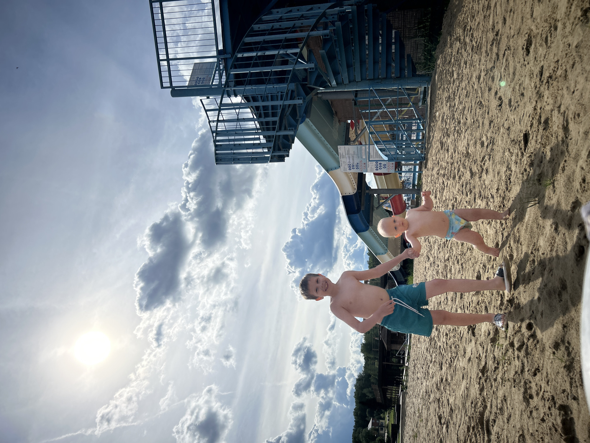 In the city of Yubileyny, Russia, two young boys are walking on a sandy beach. The older boy, wearing teal swim shorts, is holding the hand of the younger boy, who is wearing a diaper. Behind them, a blue metal spiral staircase leads up to a water slide. The sky is filled with dramatic clouds, and the sun is shining brightly. In the background, there are trees and buildings.