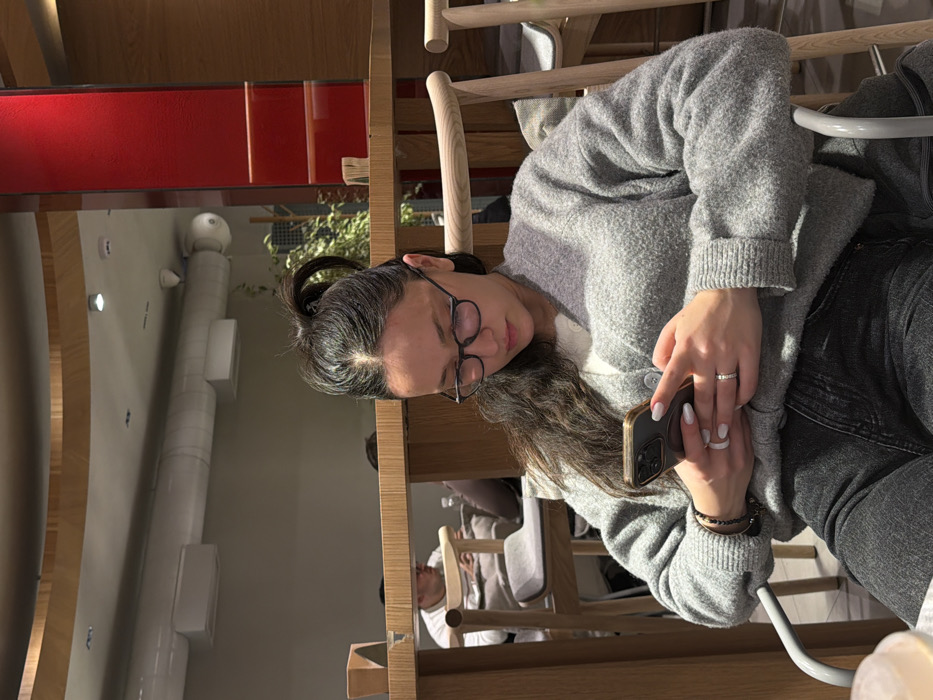 A woman with dark hair pulled back in a messy bun is seated at a wooden chair and table. She wears glasses, a gray cardigan, and dark jeans. She holds a gold phone with a black case in her hands. Her fingernails are painted a light color. In the background, other people are seated at tables. Red accent panels are visible. The background features a wall with white rectangular fixtures and a cylindrical white column.