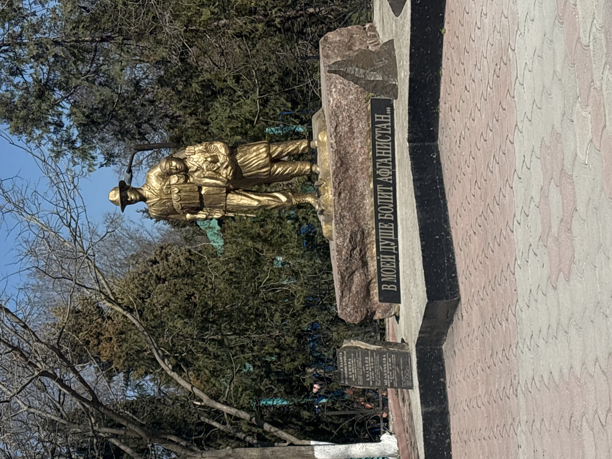 The image shows a golden-colored monument depicting two soldiers in a park-like setting with trees. One soldier appears to be carrying another. A plaque is visible with text in Russian, including the words "В МОЕЙ ДУШЕ БОЛИТ АФГАНИСТАН...", which translates to "Afghanistan hurts in my soul...". The monument is located in Chornomorskoe, Ukraine. The immediate surrounding area includes a paved surface with a decorative pattern and other memorial plaques.