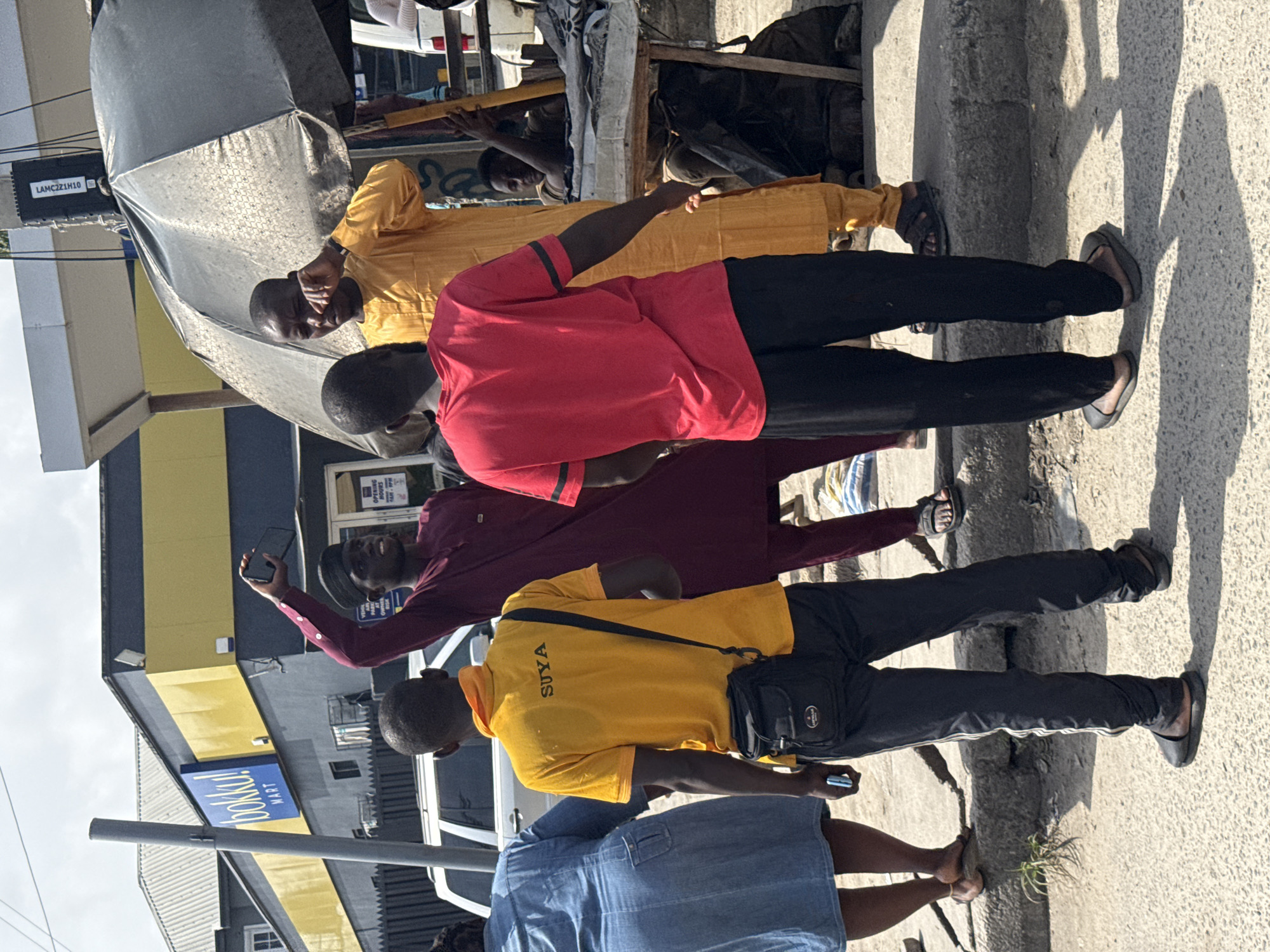 The image shows a street scene in Iranla, Nigeria, during daylight. Four dark-skinned men are walking along a sidewalk adjacent to a building, which is a branch of the Bokki Mart store. The man in front is wearing a mustard-yellow long sleeve shirt and black pants. The next man is wearing a red short sleeve shirt and black pants. Behind him, a man in a maroon long sleeve shirt is looking at his phone, and the man at the end of the line has on a mustard-yellow short sleeve shirt printed with the word "SUYA" in black capital letters, black pants, and a black bag. All men are wearing sandals. The sidewalk is concrete and in a state of disrepair with tufts of grass growing out of cracks. A black umbrella and other miscellaneous objects are on the side of the road behind the group of men.
