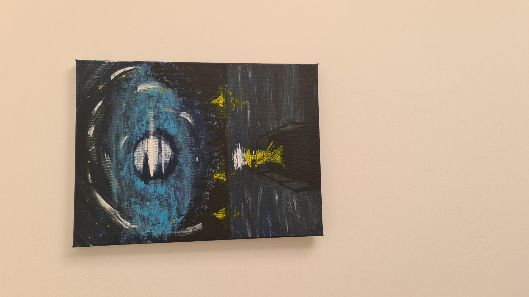 An abstract painting hangs on a light beige wall. The painting is a square canvas divided into two distinct sections. The left side features a blue and black abstract representation, possibly resembling an eye or a whirlpool. The right side is predominantly black with suggestions of building or architectural structures in dark grey. There are yellow accents scattered across both sections of the painting. The location, Budva, Montenegro, is not evident from the image. There are no people visible.
