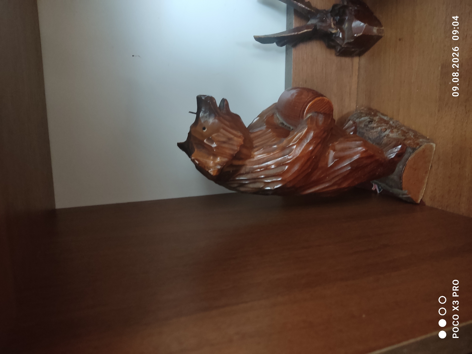 The image shows a wooden figurine of a bear on a shelf. The bear figurine is dark brown and appears to be carved from wood. It is standing on a log base and is holding a round object in its paws. The background is a plain white wall. To the right of the bear figurine, there is another wooden figurine of an eagle. The image is dated 09.08.2026, 09:04. The location is identified as Cherniakhiv, Ukraine.