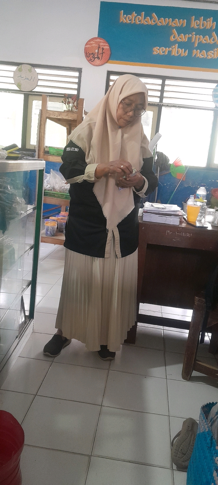 A woman wearing a light beige hijab, glasses, a black and beige long-sleeved blouse, and a long beige pleated skirt stands in a room with tiled flooring. She is holding money in her hands and appears to be counting it. Behind her, a wooden desk is visible with stacks of paper, a glass of orange liquid, a bottle of water, and a broom with green and orange bristles. The walls are white, with a blue mural at the top featuring yellow text and two circular decorations with Arabic calligraphy. A glass display cabinet is to the left of the woman, and a wooden chair is to the right of the desk. The lighting suggests an indoor setting, possibly a classroom or office.