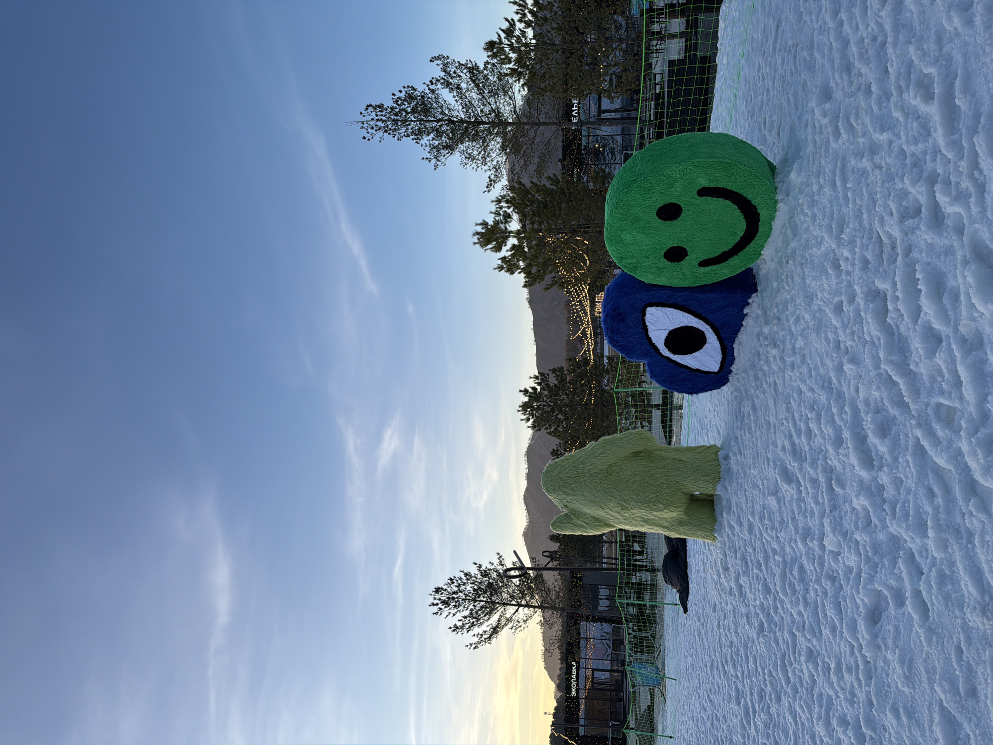 The image shows a snowy outdoor scene in Озёрное, Russia. A green smiley face and a blue monster-like plush toy are stacked on the snow, near a green fence. In the background, there are trees, a building with lights, and mountains under a blue sky.