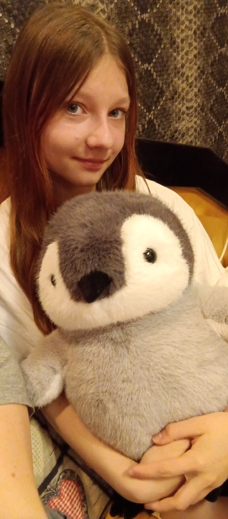 A medium close-up portrait shows a young female holding a large, plush penguin. The girl, positioned slightly to the left of the frame, has long, reddish-brown hair parted down the middle and blue eyes, looking towards the camera with a gentle, soft smile. She wears a white top, with a grey sleeve visible on her left arm, and holds the stuffed animal with both hands.
The soft toy is a grey and white penguin, with a dark grey head featuring white markings around its black eyes and a black beak. It occupies the central and lower part of the frame.
The background behind the girl's head and left shoulder is a dark, textured wall or wallpaper with a subtle, reflective diamond-like pattern. To her right, the background includes a warm-toned, possibly wooden or upholstered surface and a darker, flat area. In the foreground, beneath the penguin, a patterned fabric featuring red and white checkered heart shapes and other designs is visible. The scene is lit with soft, indoor lighting. There is no visible text in the image. The setting appears to be an indoor environment.