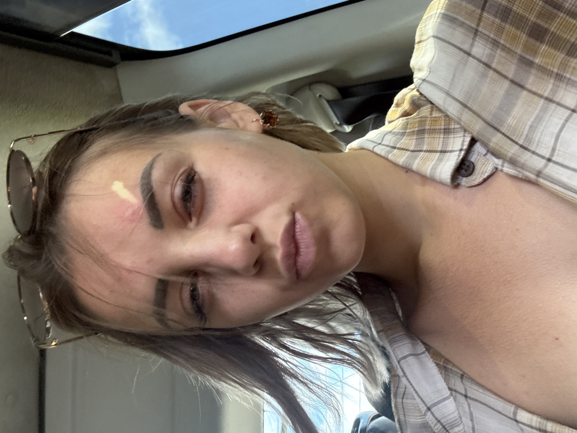 A young woman with blonde hair pulled back into a ponytail is taking a selfie in a car. She is wearing a plaid shirt and sunglasses on her head. Her hair is blowing in the wind. The sky is visible through the car window. There is a bright spot on her forehead, possibly from the sun.