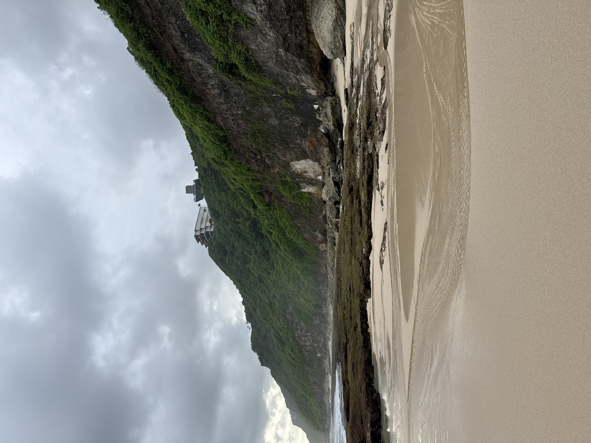 The image captures a coastal scene in Pecatu, Indonesia, featuring a cliff face covered in green vegetation. A multi-story building sits atop the cliff. Below the cliff, there is a beach with sand, and shallow water reflecting patterns. The sky is overcast with gray clouds. No people are visible in the frame.